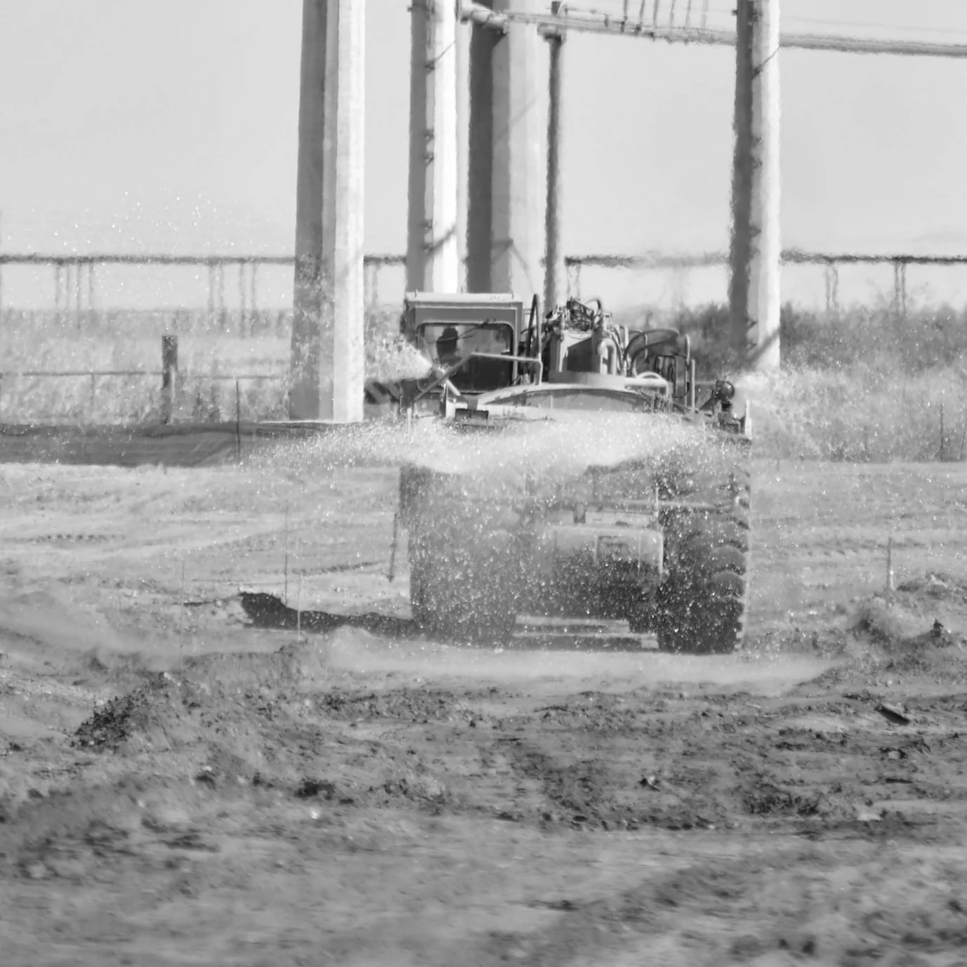 A monochrome image of a construction scene with a water truck spraying water on a dirt road, with pillars in the background.