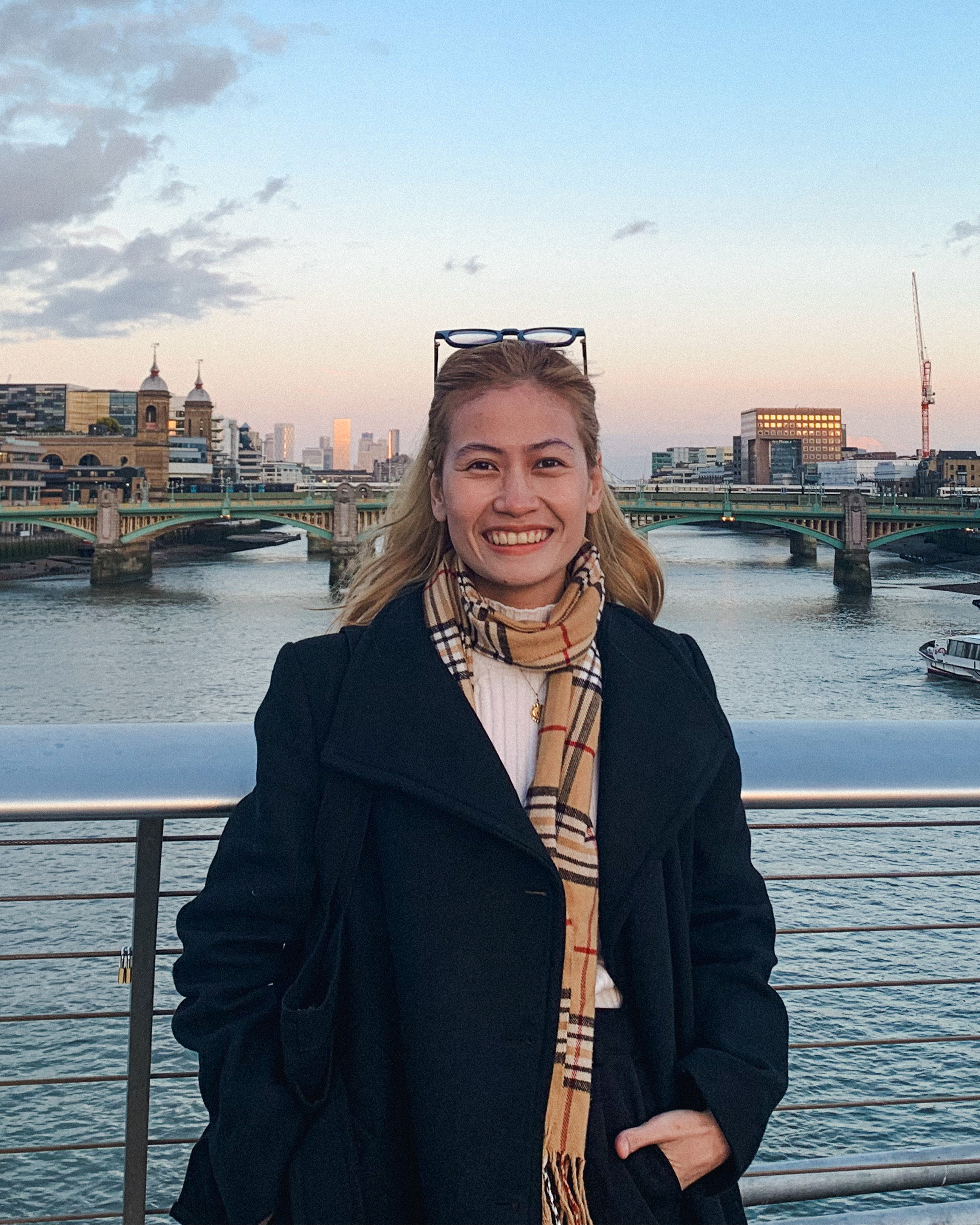 A woman smiling on a bridge over a river with city buildings and a bridge in the background during sunset.