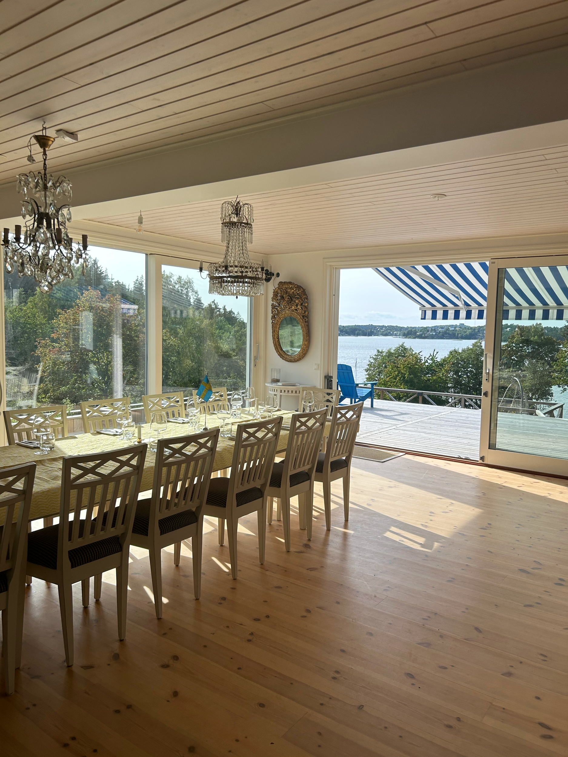 A dining room with a wooden floor, long table, multiple beige chairs, large windows showing an outdoor patio with a blue chair and water in the background, and chandeliers hanging from the ceiling.