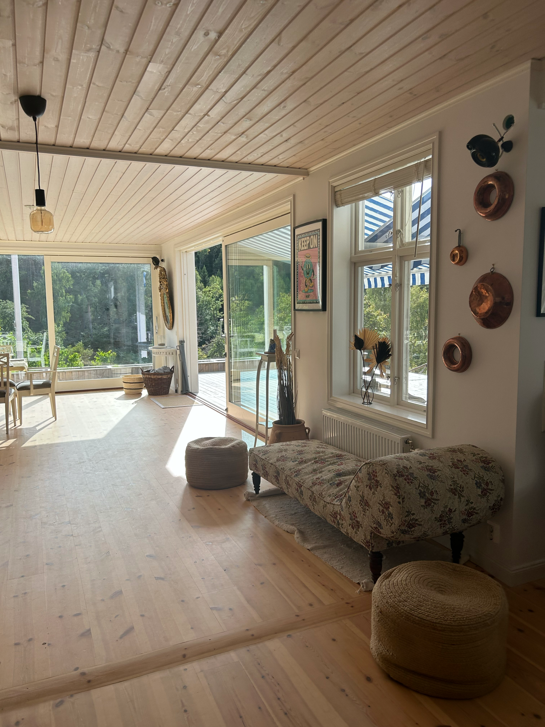 Living room with wooden wall paneling, floral sofa, round woven pouf, artwork, and large windows with views of greenery and outdoor patio.