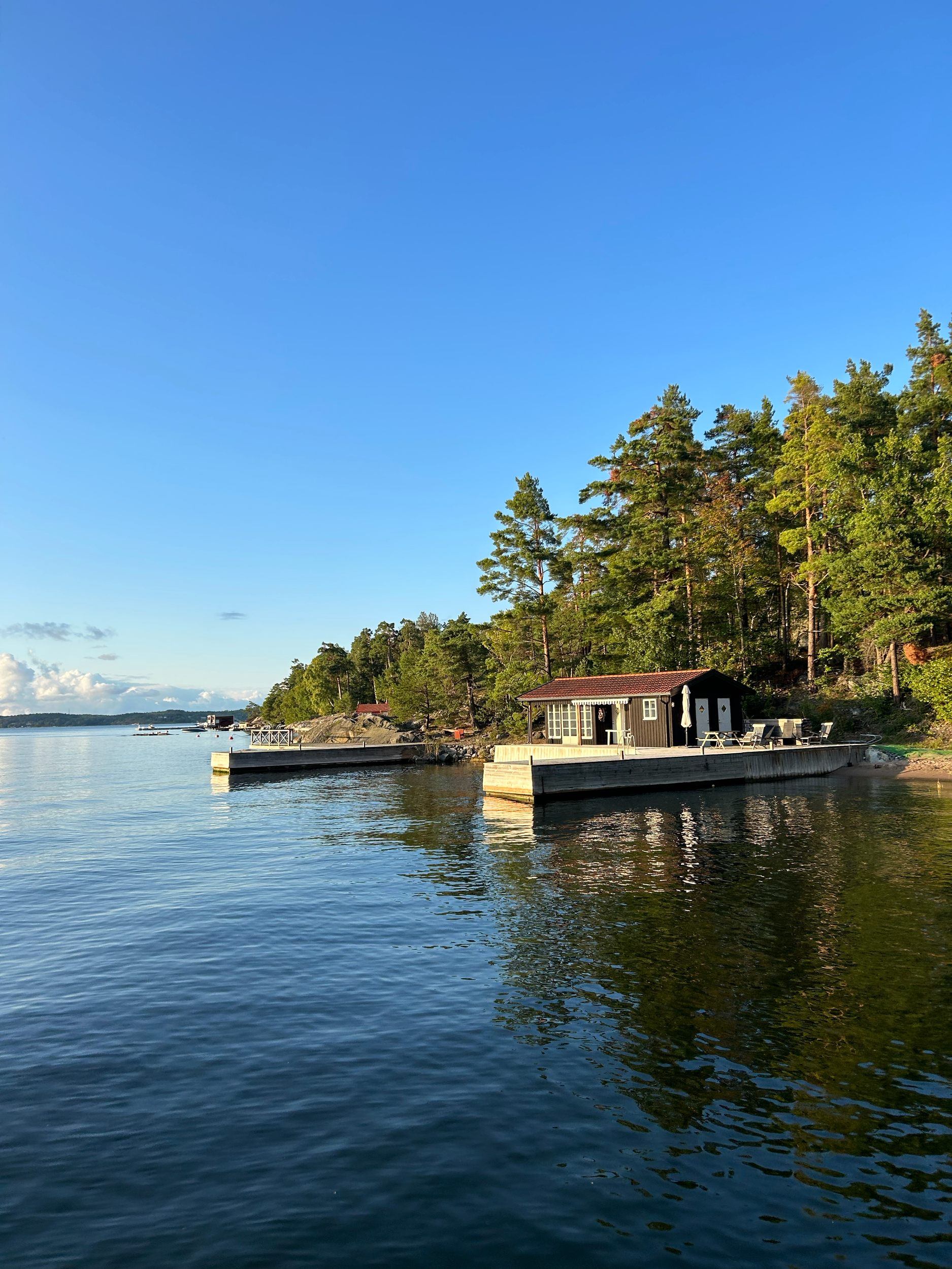 A lakeside house surrounded by trees, with a small dock extending into the water and a boat docked nearby, under a clear blue sky.