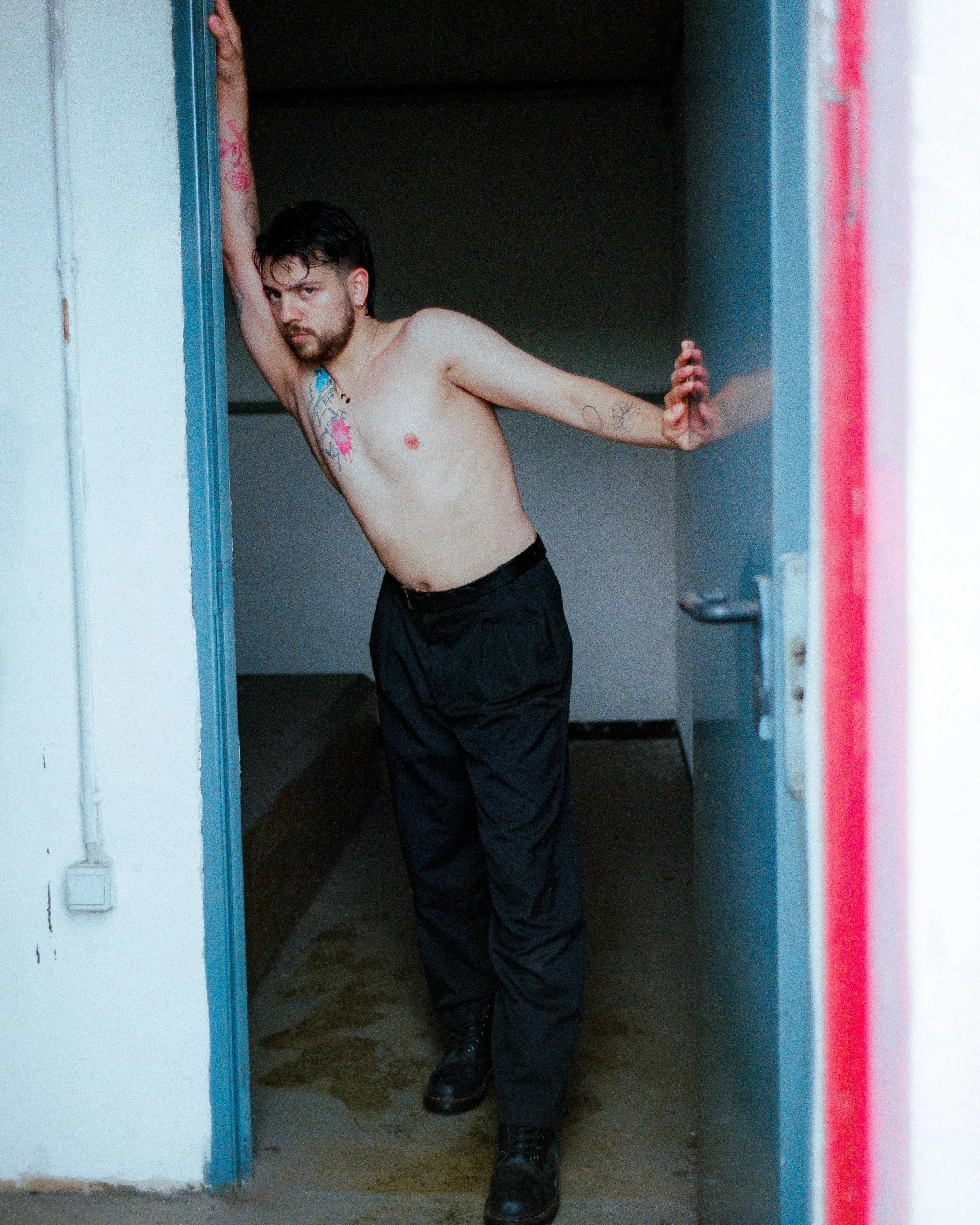 A shirtless man with tattoos standing in a doorway, leaning to one side and looking at the camera.