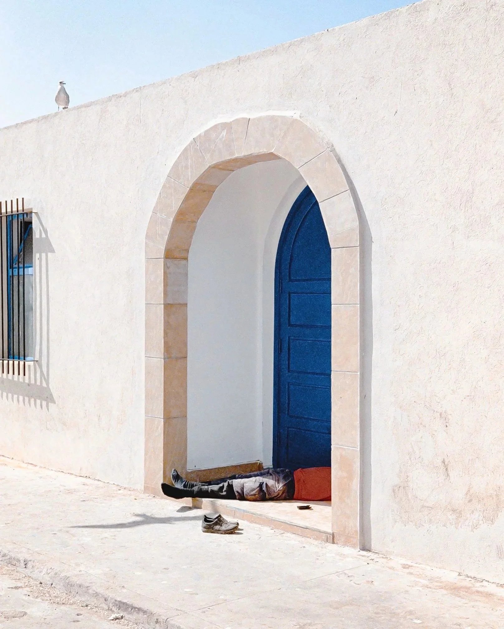 A person lying on a ledge in front of a blue door with an arched stone frame, on a white wall, with a bird perched on the wall above.