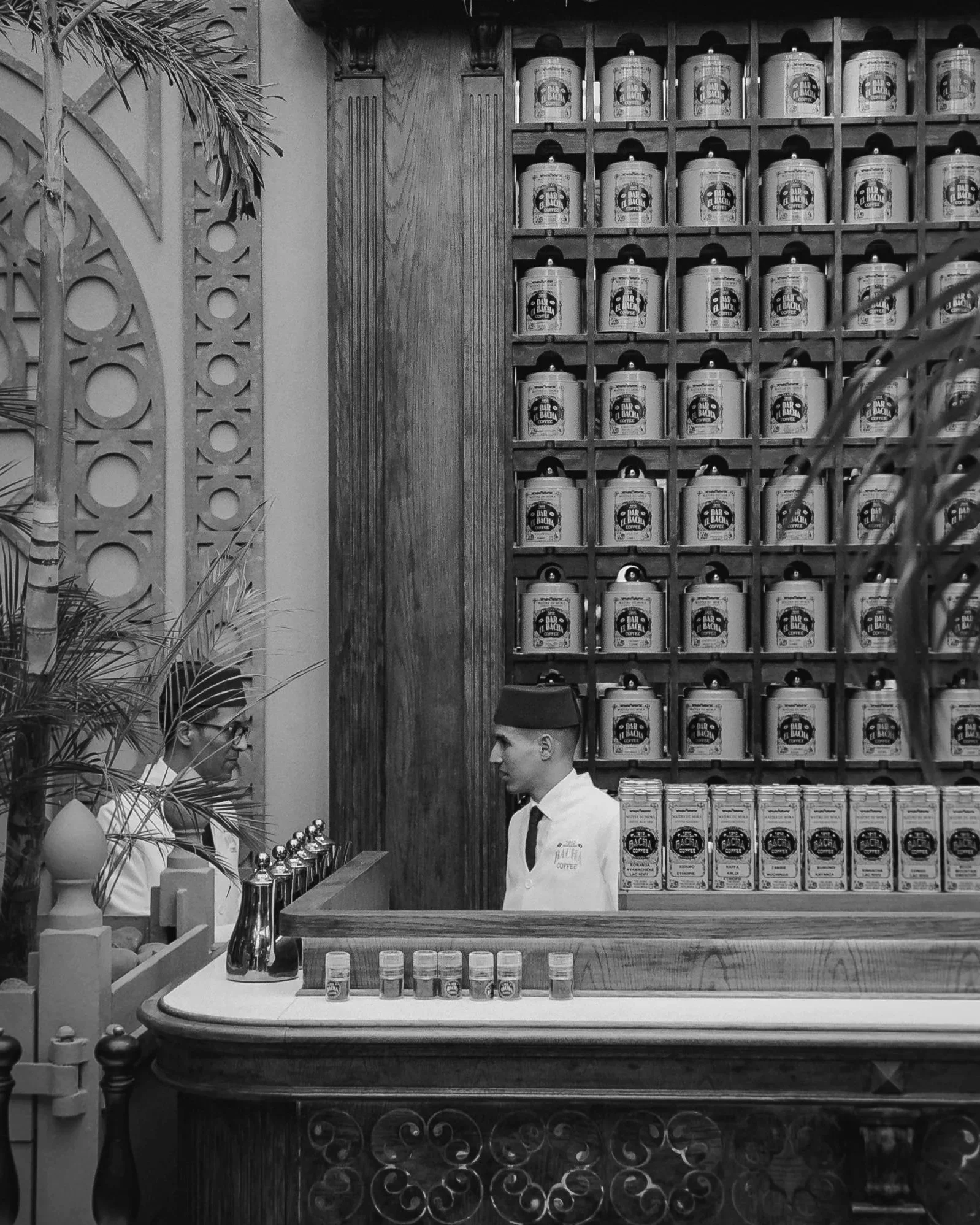 A vintage-style coffee shop interior with a wooden counter, a wall of labeled coffee containers, and two baristas in uniform. One barista wears a fez, the other glasses.