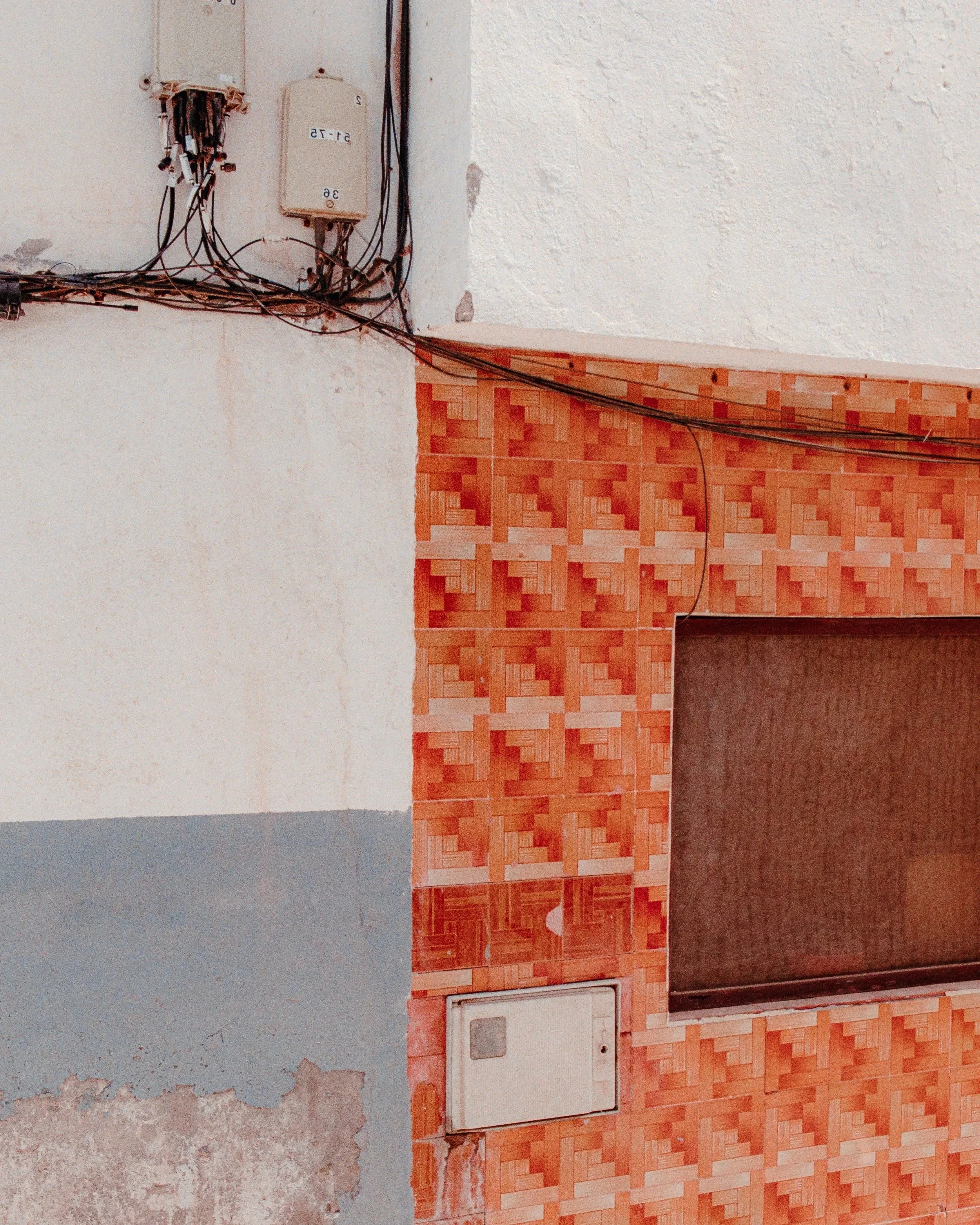 Close-up of a wall with exposed electrical wires, a small white electrical box, and a window with a red patterned tile border.
