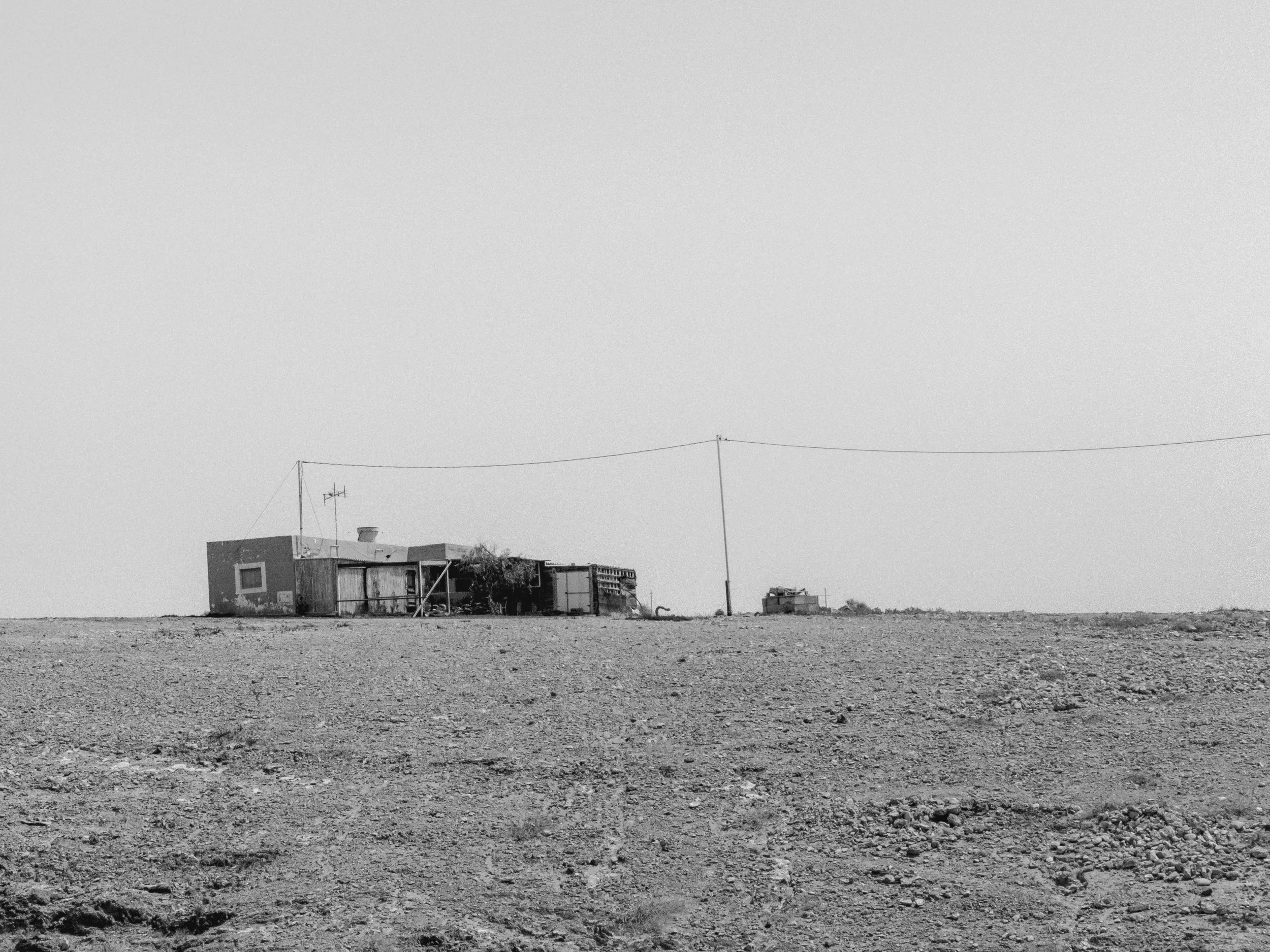 A remote, barren landscape with a small, weathered house and a utility pole connected by wires, under a vast, clear sky.