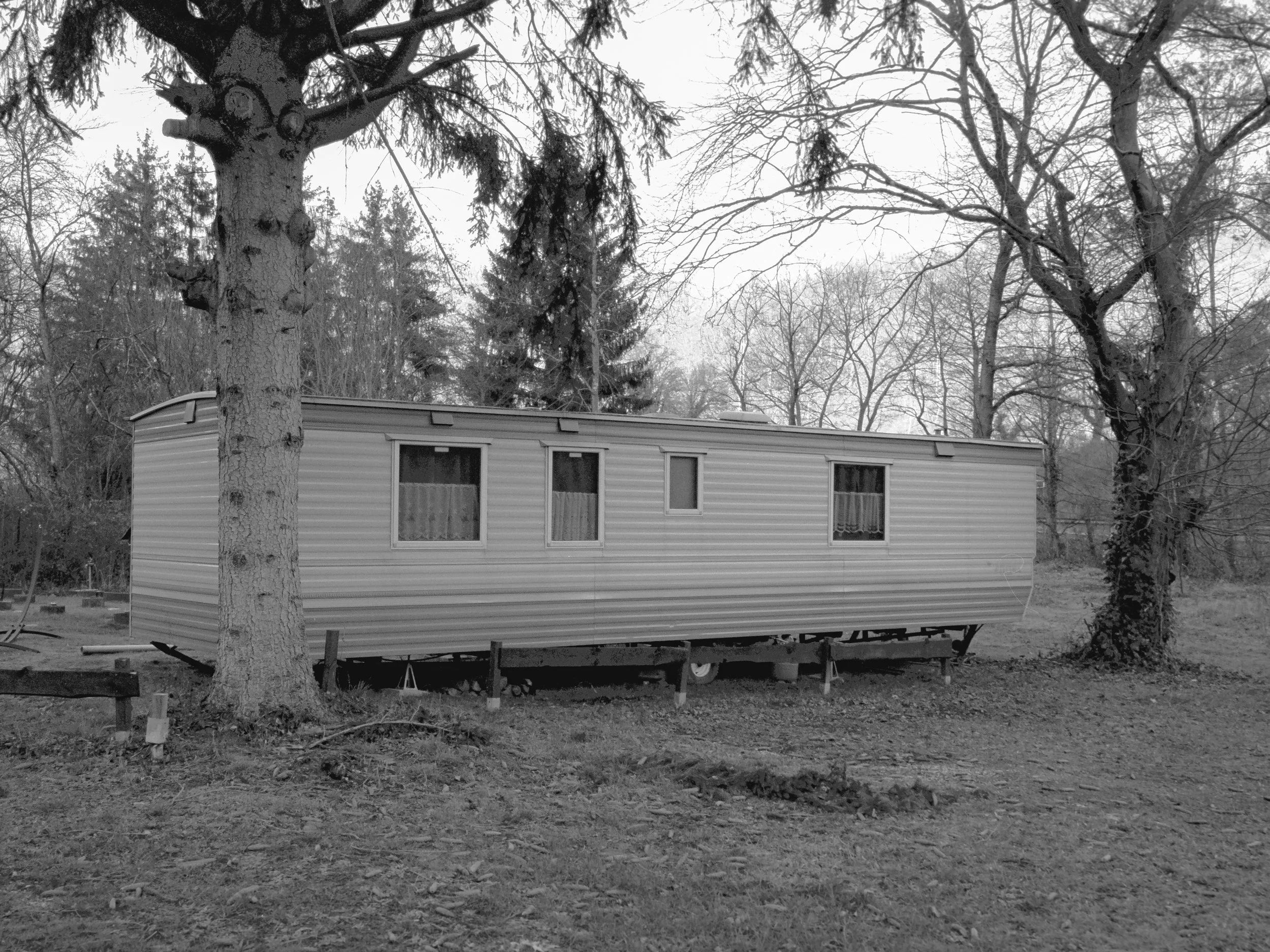 A mobile home on stilts in a wooded outdoor setting with leafless trees surrounding it.