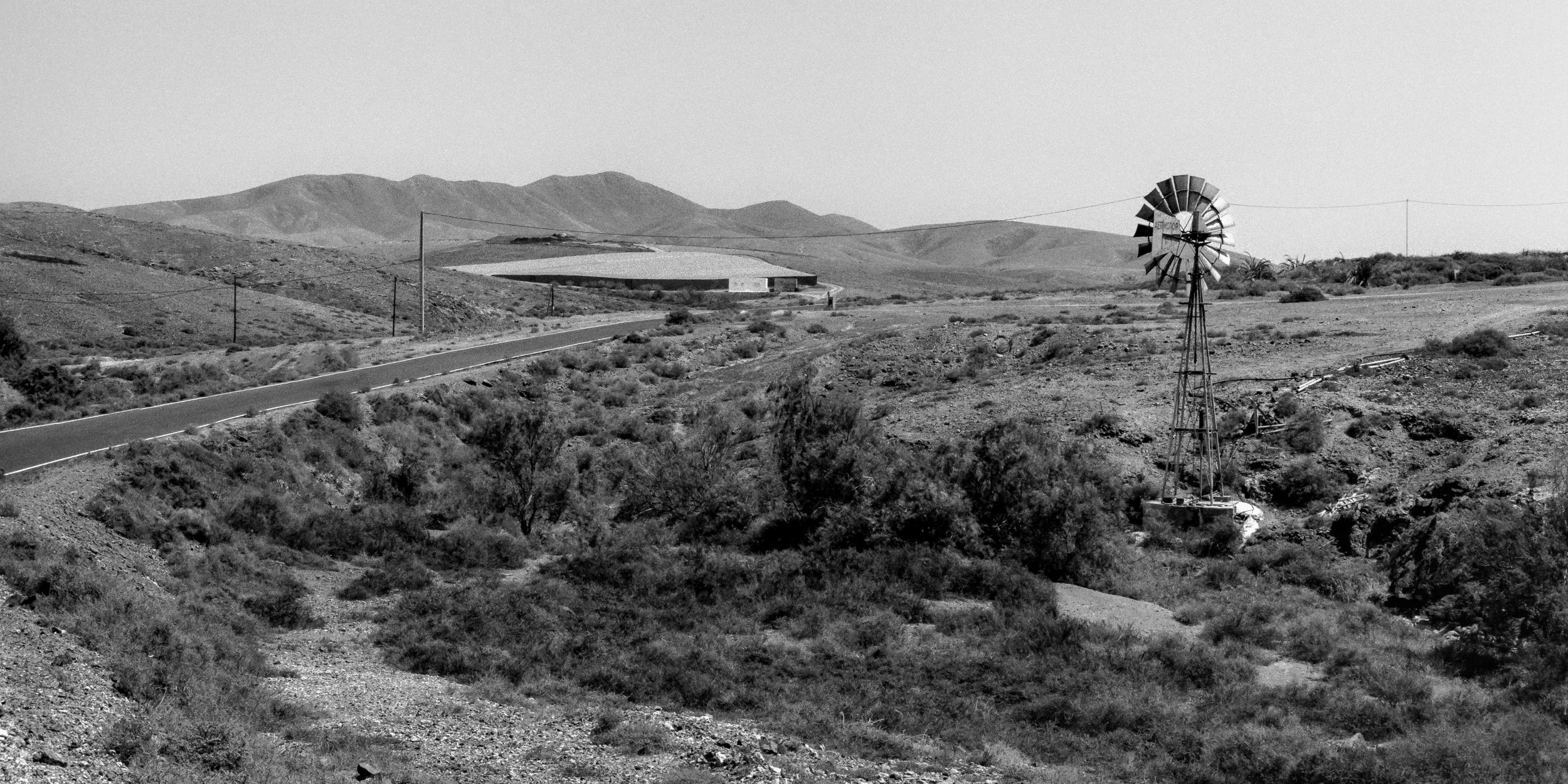 A black and white landscape of a desert area with hills in the background, a winding road, a small building, and an old windmill.
