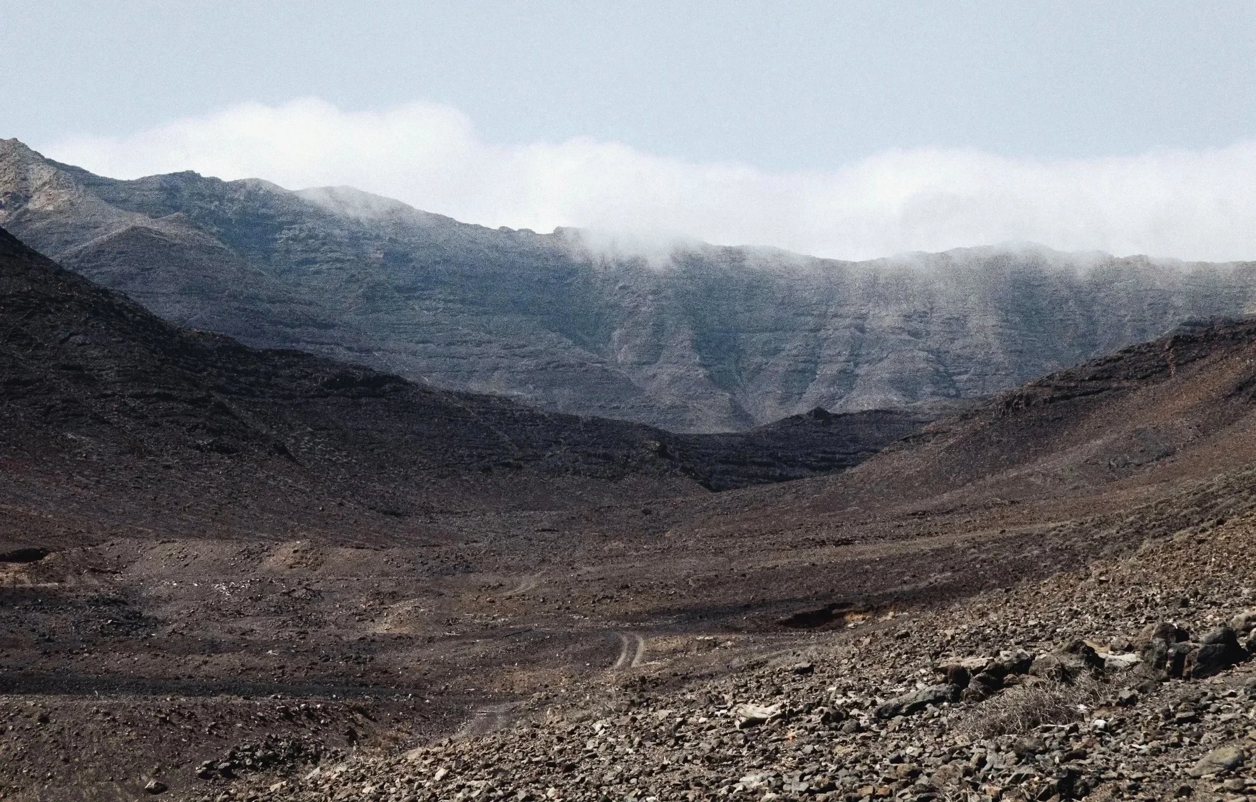 A rocky mountain landscape with dark, rugged terrain in the foreground and mountain peaks in the background, partially covered by clouds.