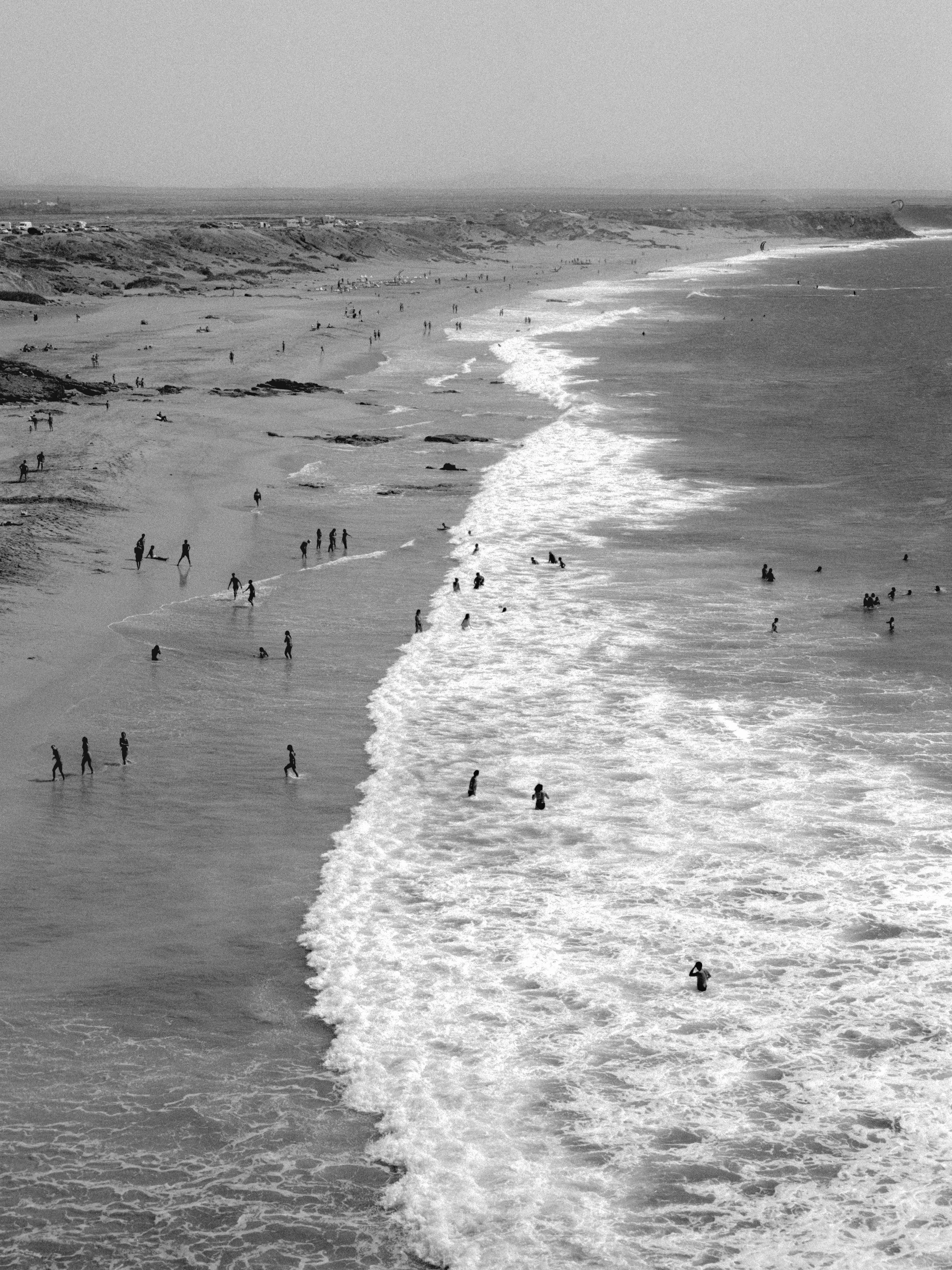 People enjoying a day at the beach, some walking along the shoreline and others swimming or playing in the water, with a scenic coastline in the background.