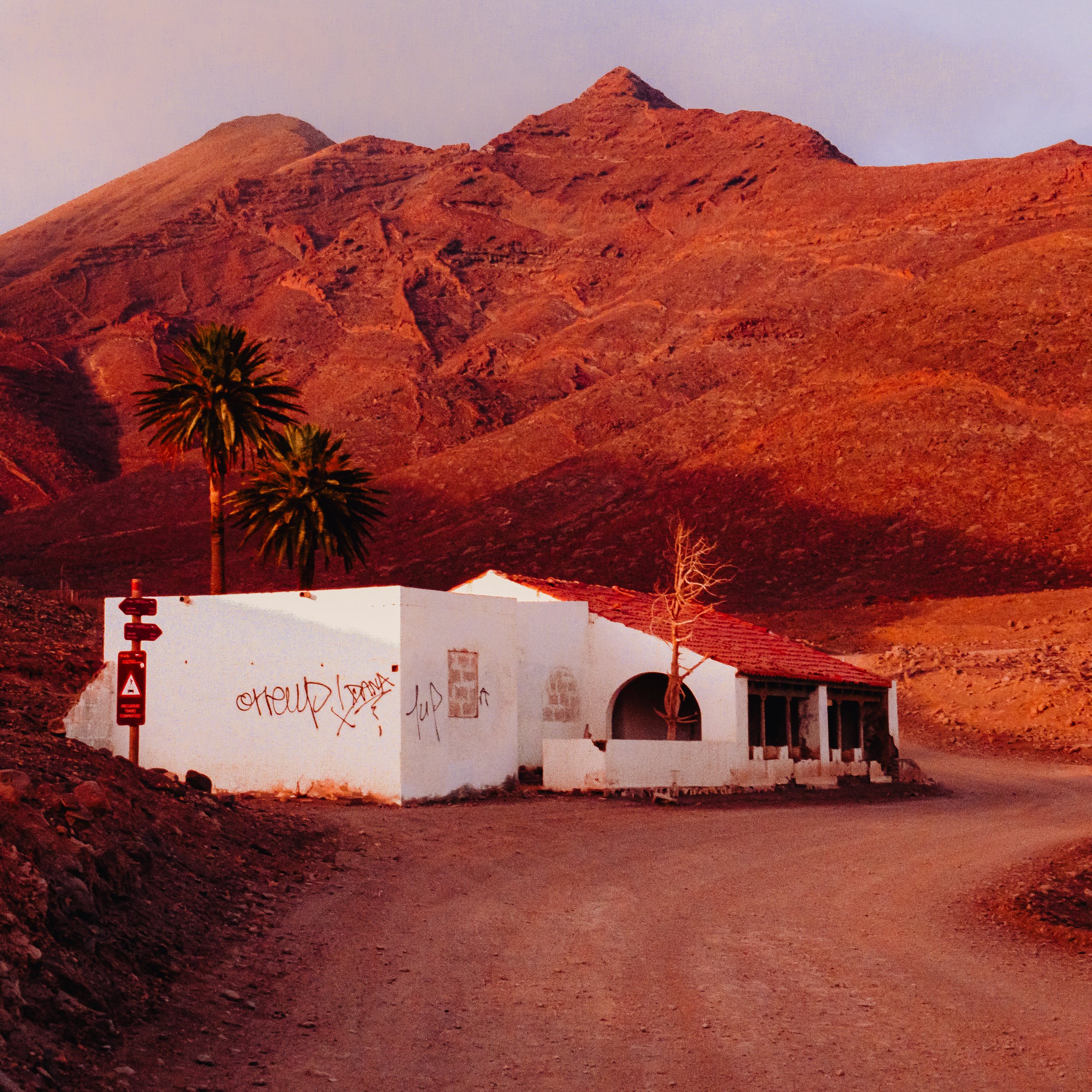 A white building with graffiti on the side, set against arid mountainous landscape with palm trees and a dirt road.