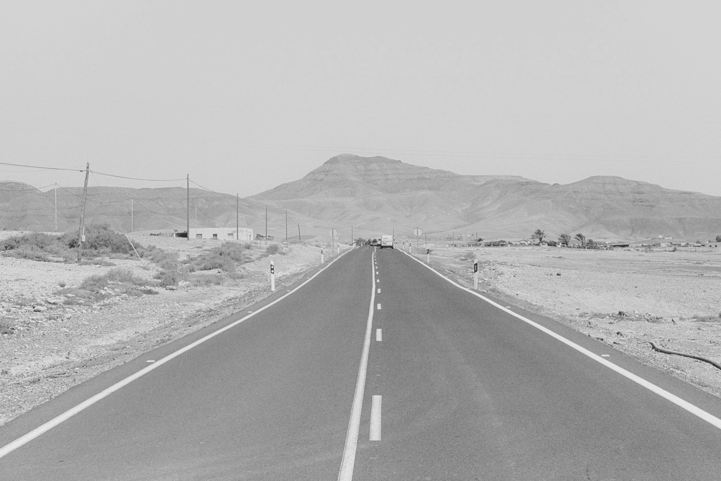 A black and white photo of a straight, empty two-lane road in a desert landscape with distant mountains and a small building on the left side.