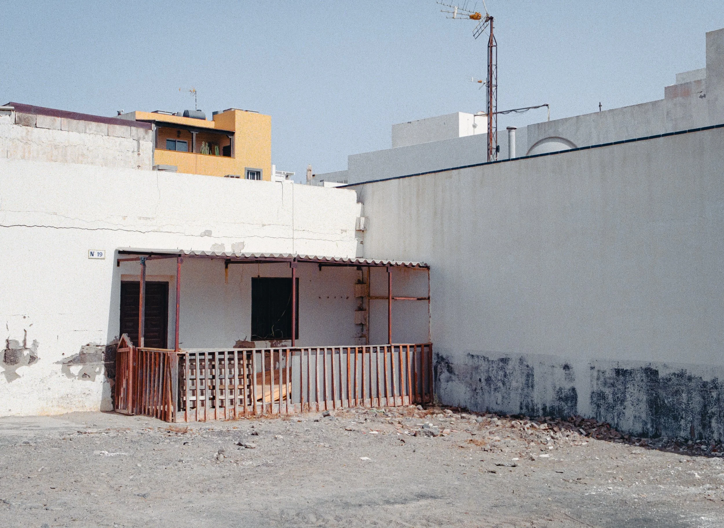 An empty lot with a mostly white building facade, a small balcony with a wooden railing, and a dirt ground with some debris. In the background, there are neighboring buildings and antennas on rooftops.