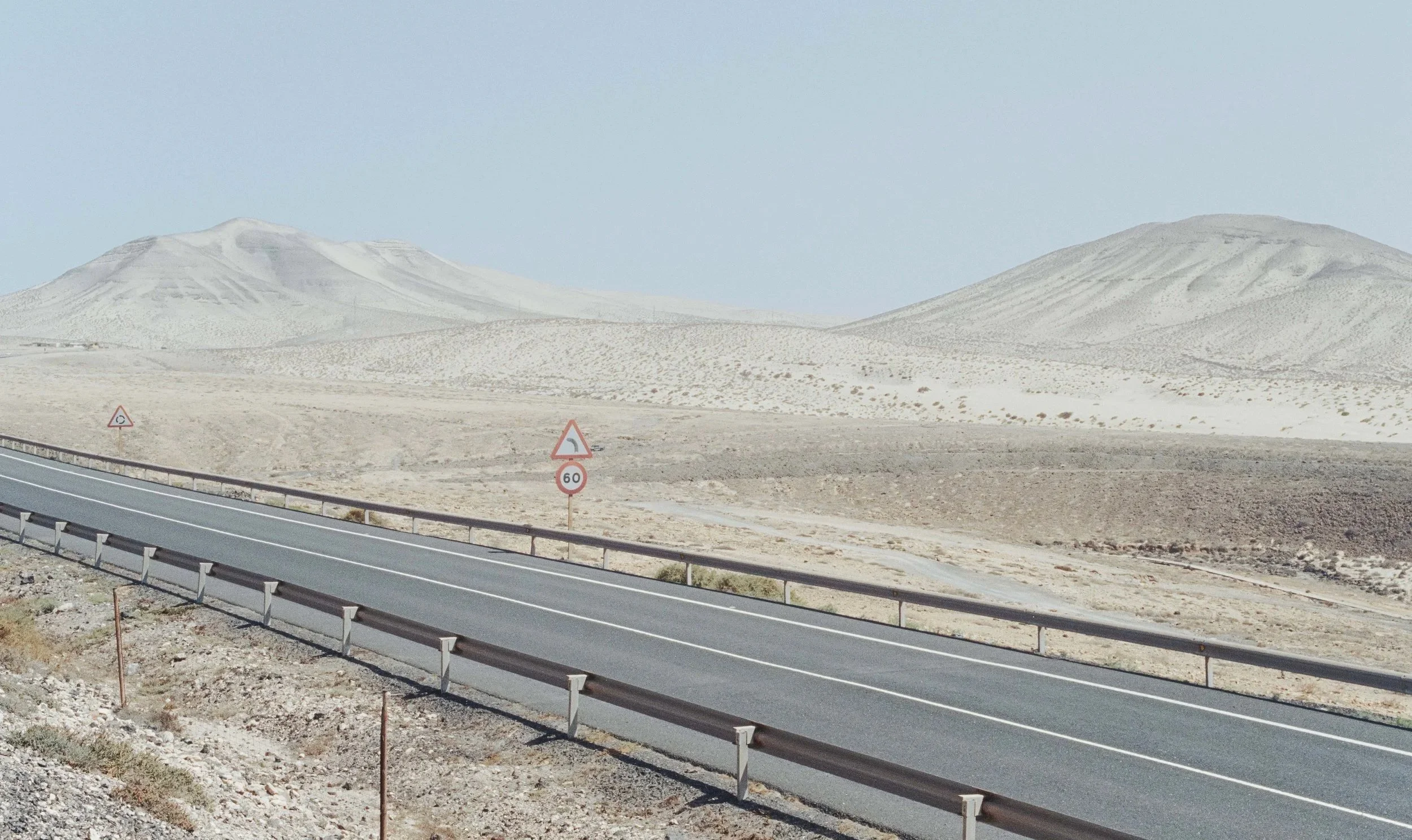 A road running through a desert landscape with sand dunes in the background. There are road signs along the road indicating a curve and a 60 km/h speed limit.