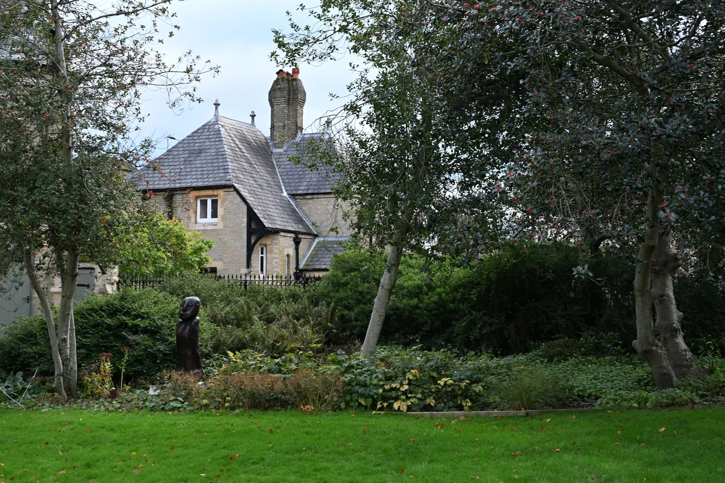 A garden with green grass, various trees, bushes, and a black sculpture of a human figure. In the background, there is an old stone house with a slate roof and chimney, surrounded by a black wrought iron fence.