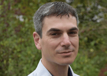 Portrait of a man with short dark hair, wearing a light blue collared shirt, outdoors with trees in the background.