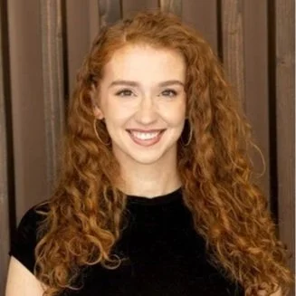 A young woman with curly red hair and a black shirt, smiling in front of a wooden background.