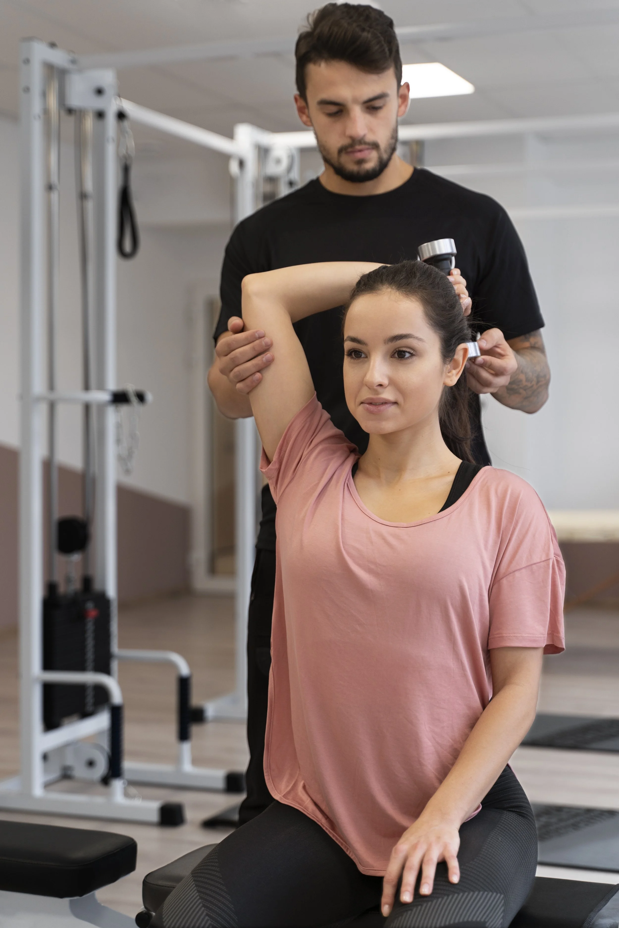 A young woman is doing a stretching exercise while seated on a black exercise mat in a gym, with a personal trainer helping her.