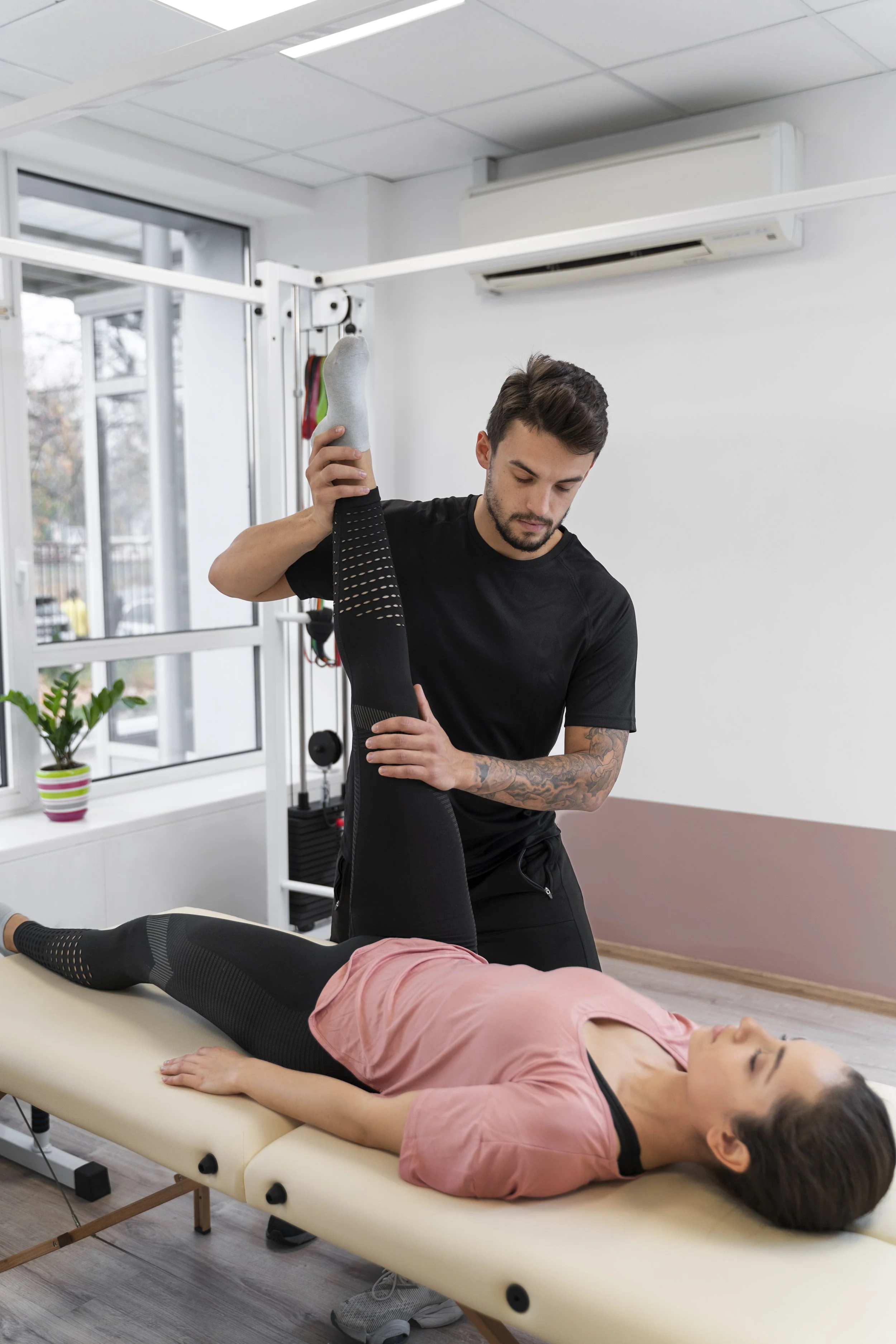 A physical therapist performing leg stretching exercises on a woman lying on a treatment table in a therapy room.