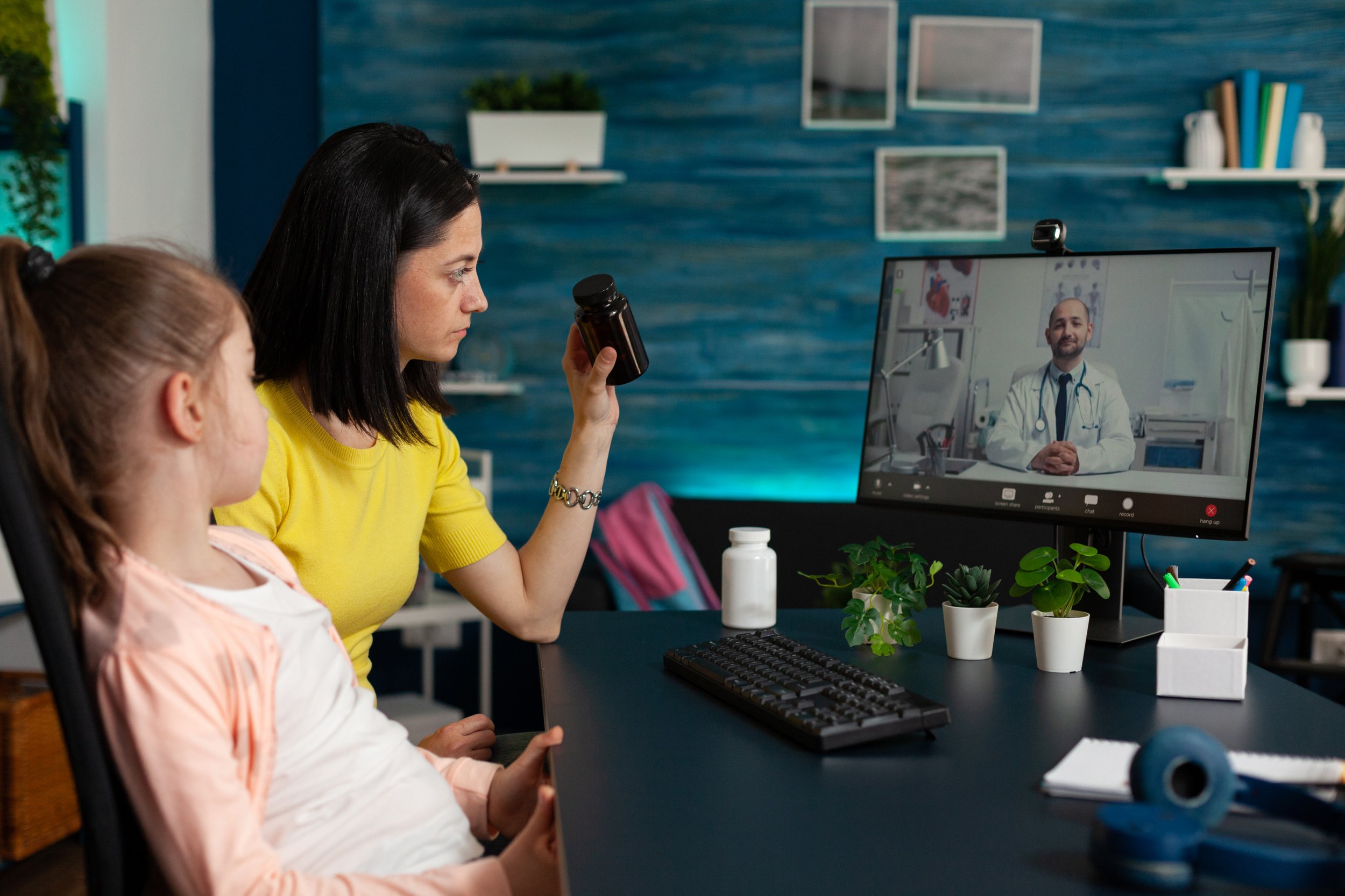 A woman and a young girl are engaging in a virtual consultation with a doctor on a computer screen. The woman is holding a bottle, possibly medication or supplement. The room has a dark desk with a keyboard, potted plants, and a computer monitor displaying a doctor in a white coat with a stethoscope.