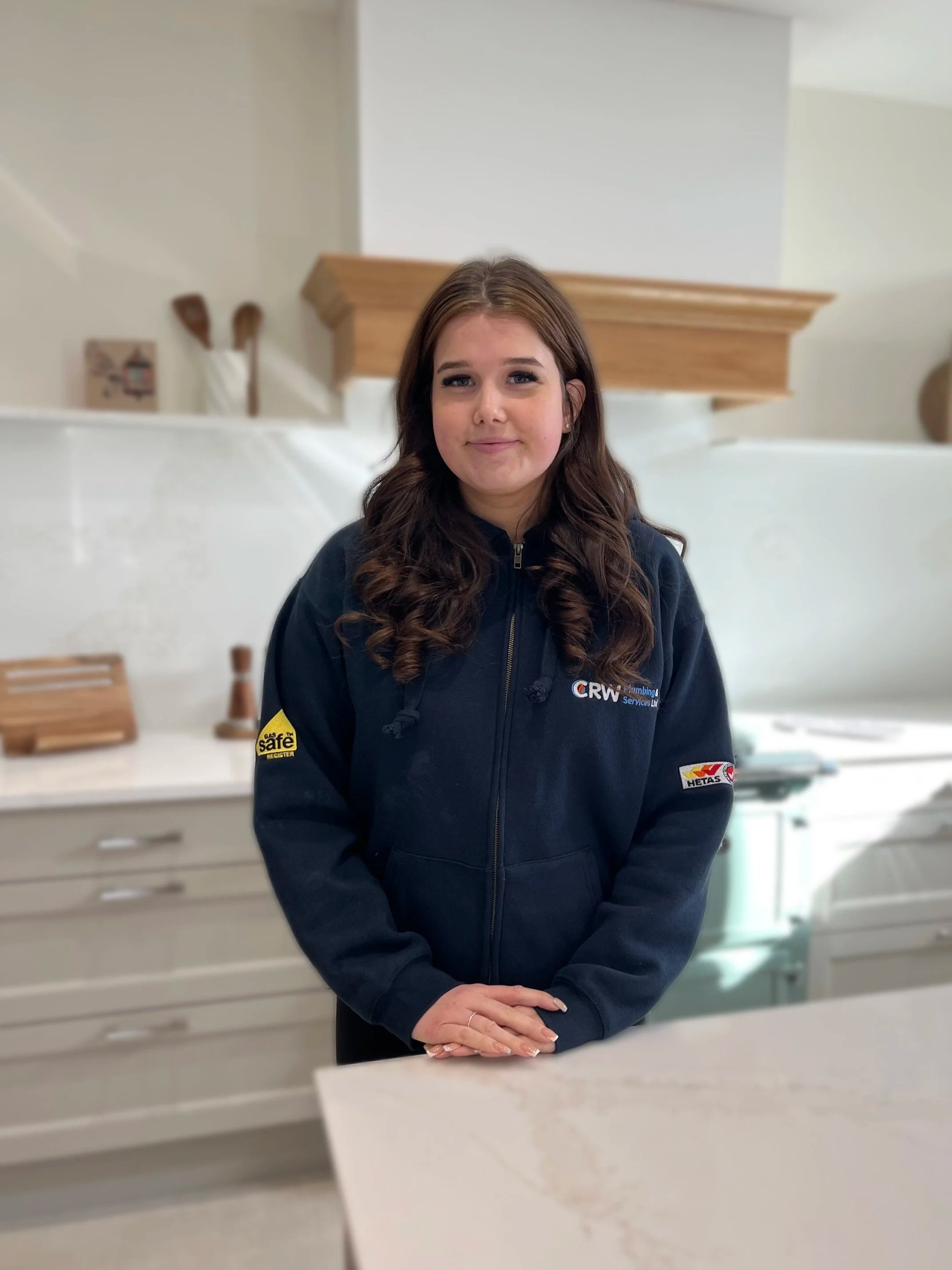 A young woman with long brown hair, wearing a navy blue polo shirt, smiling gently at the camera, standing indoors in front of a neutral background.