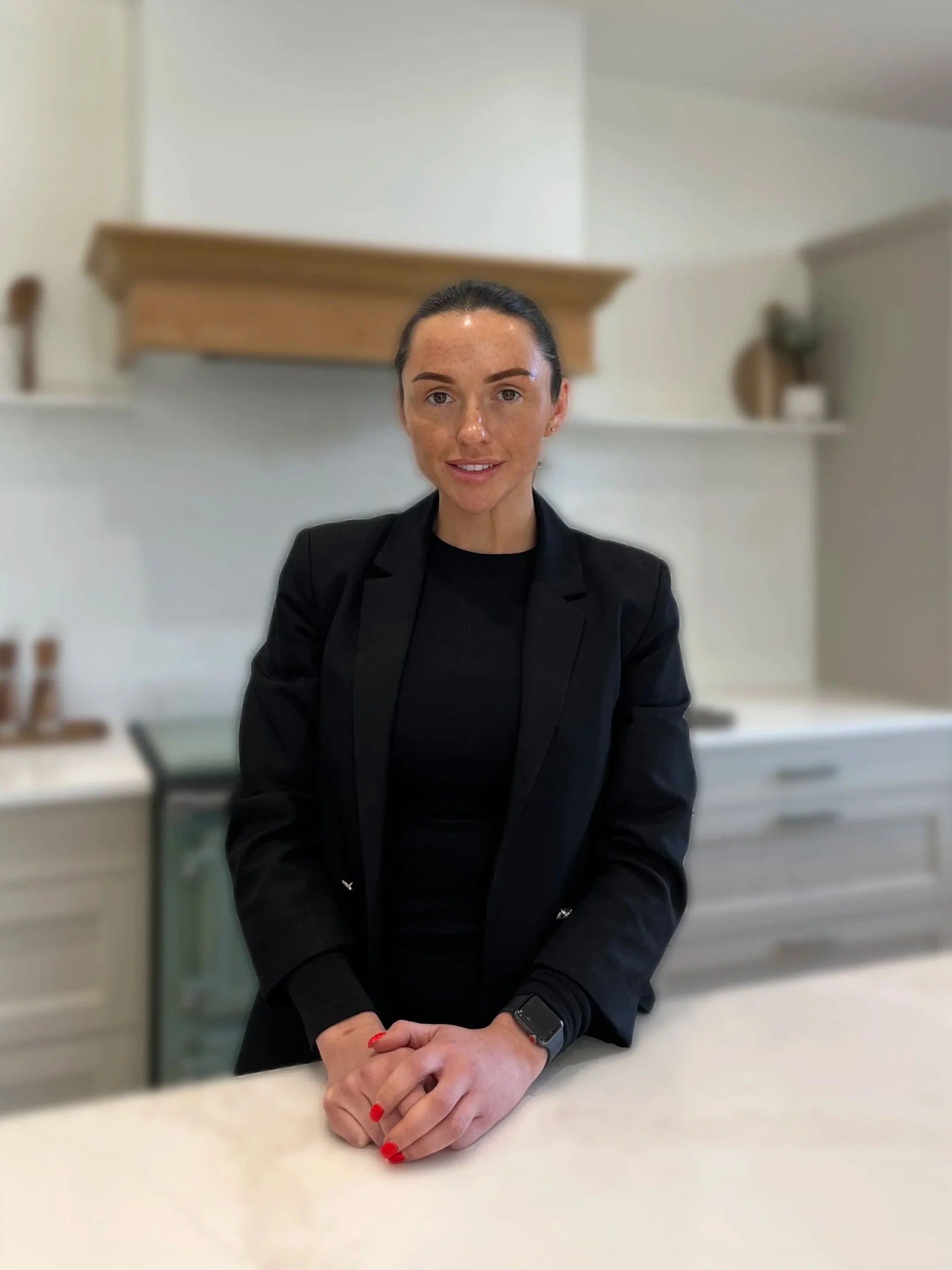 A woman with dark hair tied back, wearing a black blazer and black top, smiling in an indoor setting with stairs in the background.