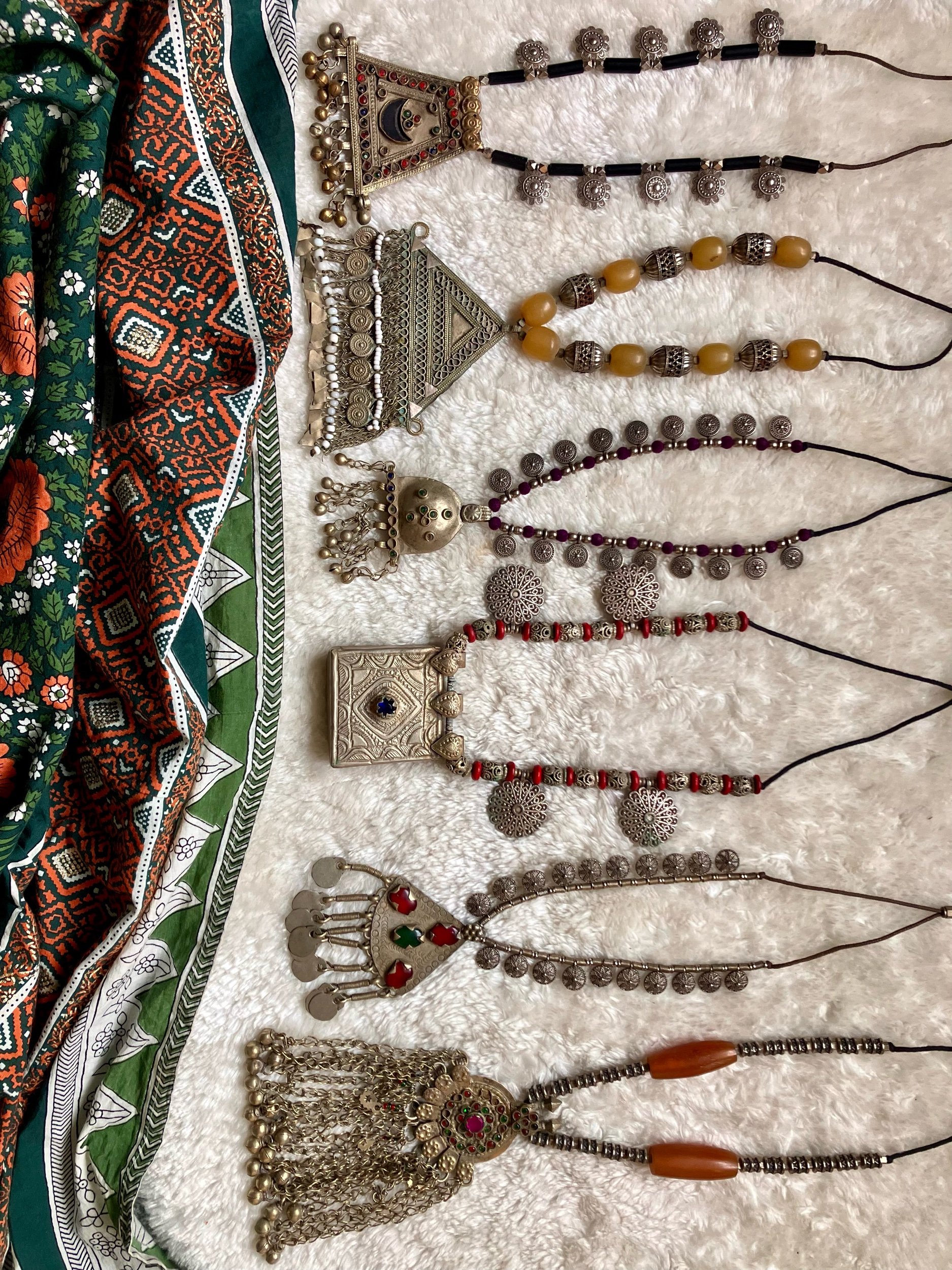 Seven decorative necklaces with traditional jewelry designs are laid out on a soft white surface next to a colorful patterned fabric with floral and geometric patterns.