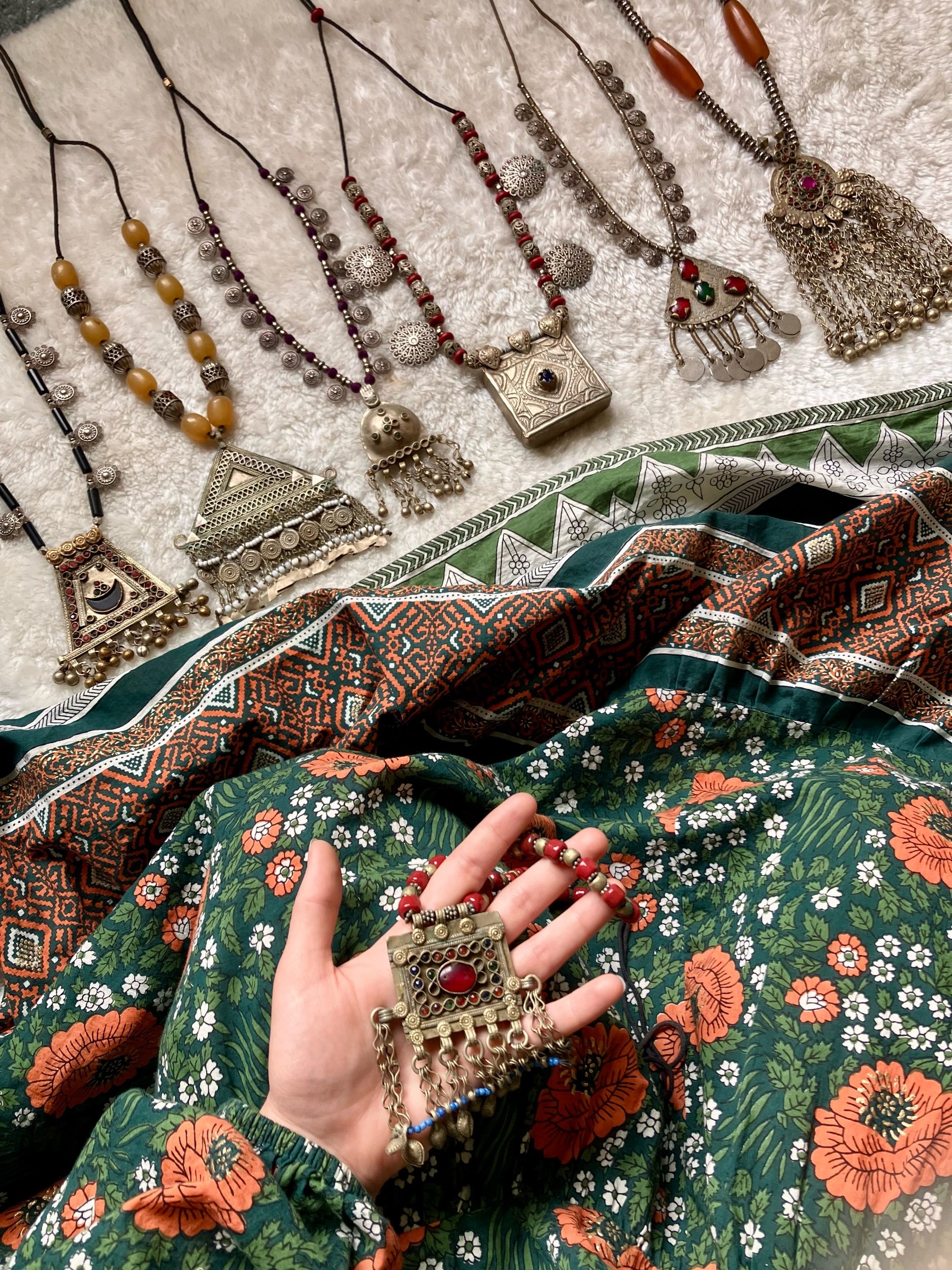 Collection of ornate jewelry including necklaces with beads and metal pendants, laid out on a textured surface, with a hand holding a large, decorative pendant necklace with a red stone and dangling charms, all set against a floral and patterned fabr