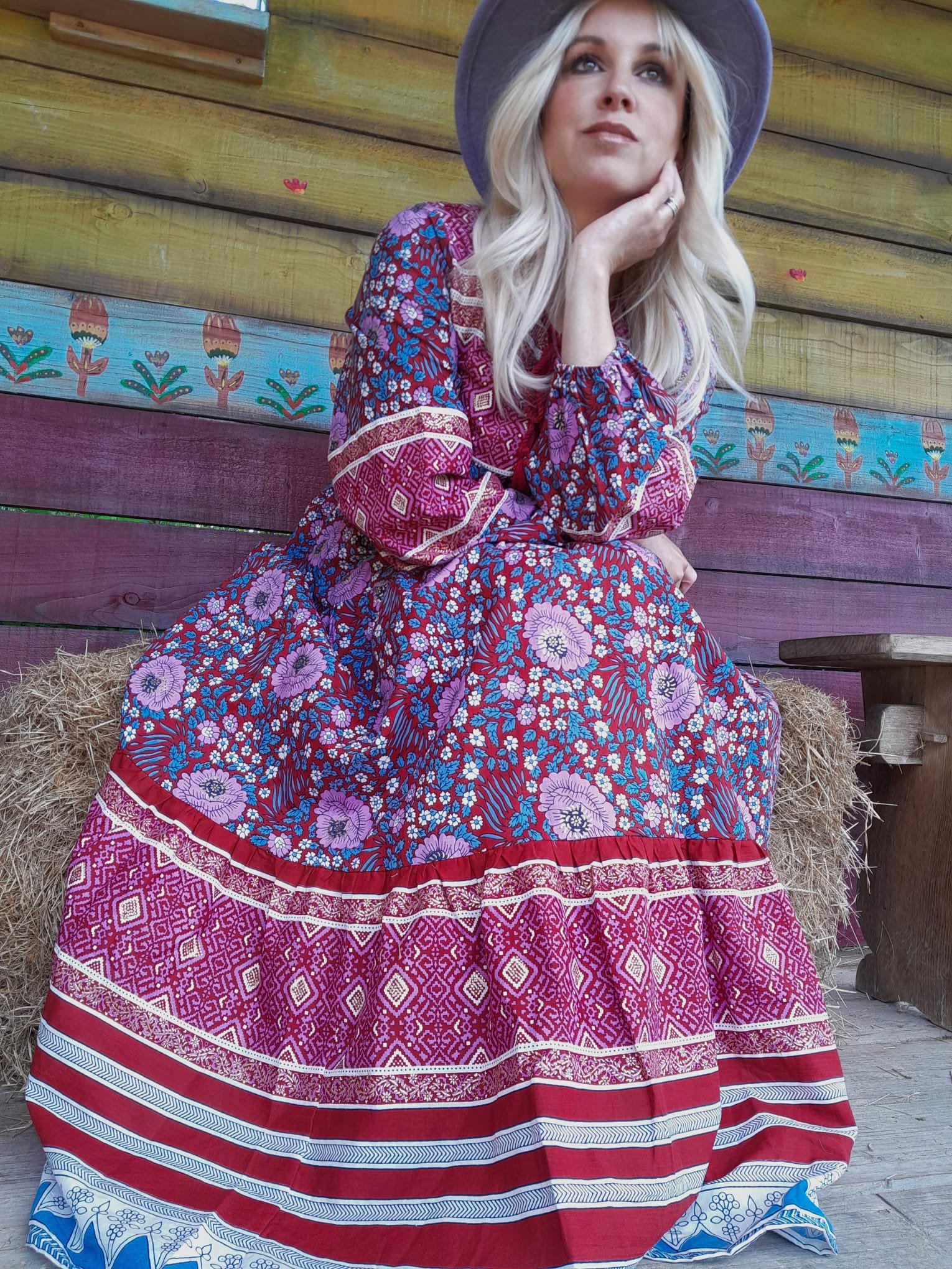 A woman with long blonde hair, wearing a purple hat and a long, colorful paisley-patterned dress, sitting on a hay bale in front of a painted wooden wall.