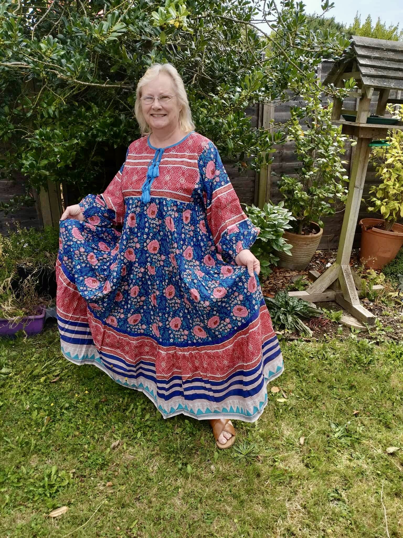 A smiling woman with blonde hair and glasses wearing a multicolored, floral, and striped dress standing outdoors on a grassy yard with potted plants and a wooden fence in the background.