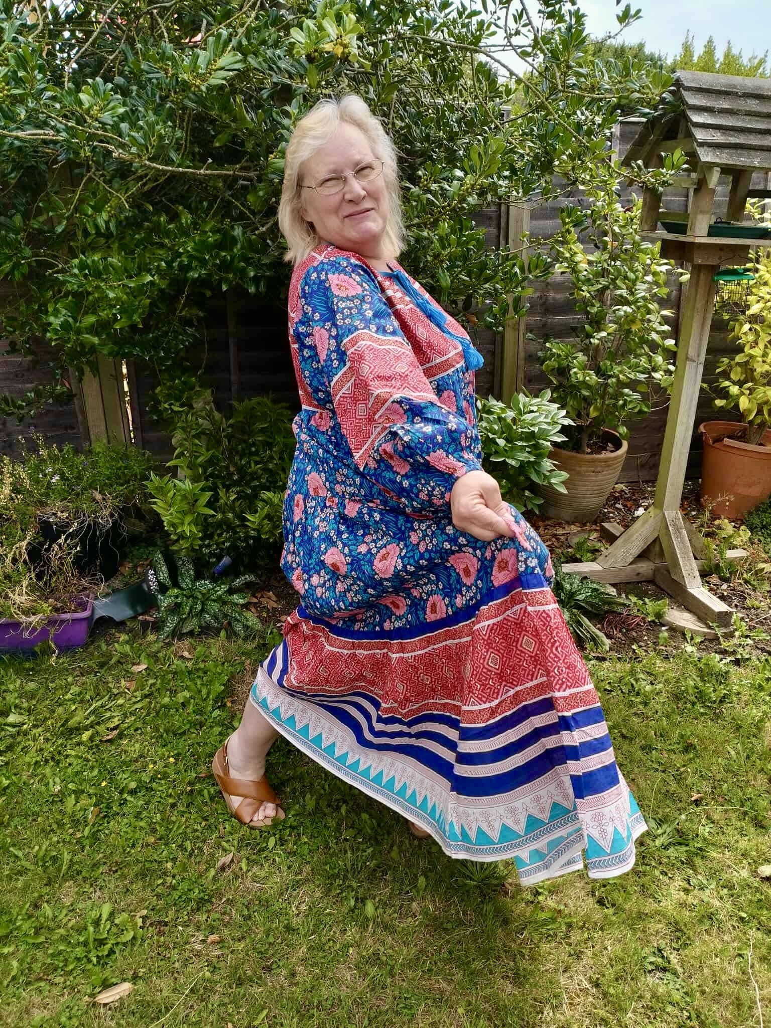 A woman with blonde hair and glasses wearing a colorful patterned dress standing outdoors in a garden with green plants, smiling and posing with a playful stance.
