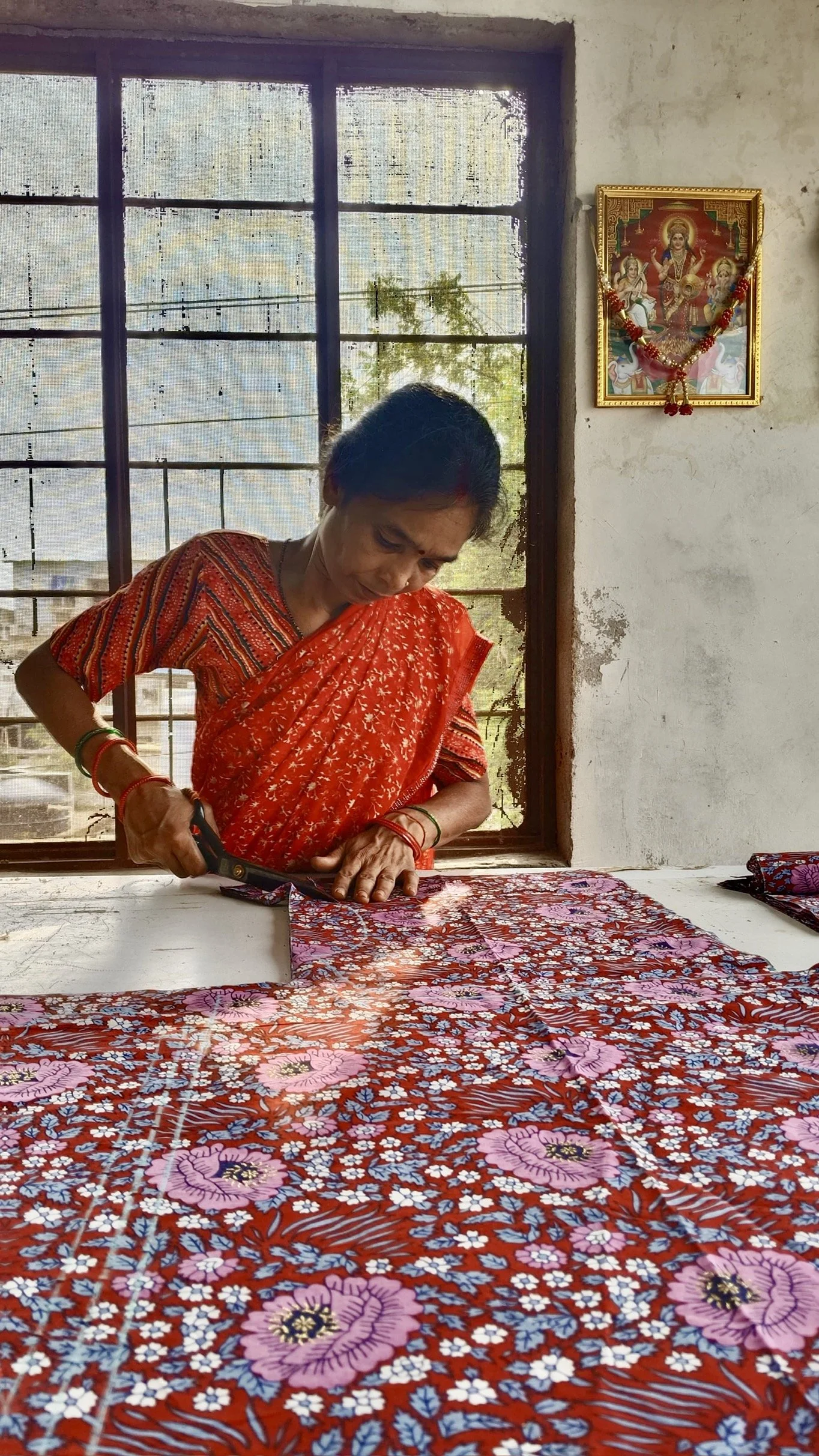 An elderly woman dressed in a red patterned sari is cutting a colorful floral fabric on a white table inside a room with a large window behind her. There is a Hindu religious picture on the wall decorated with a garland.