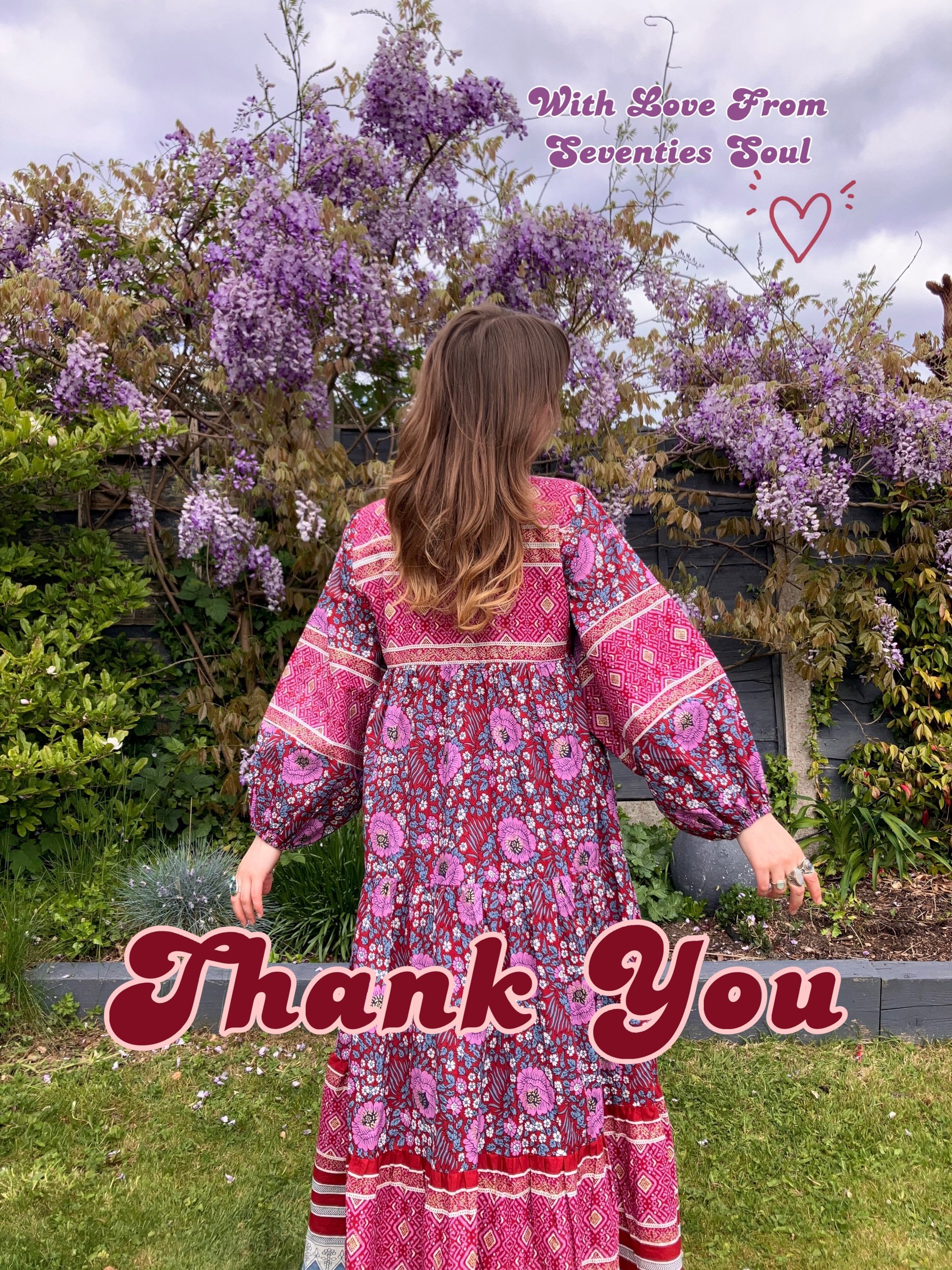 A woman with long wavy brown hair wearing a colorful bohemian-style dress stands outdoors in a garden with purple blooming wisteria and other green plants, with an overcast sky in the background, surrounded by thank you and love message overlays.