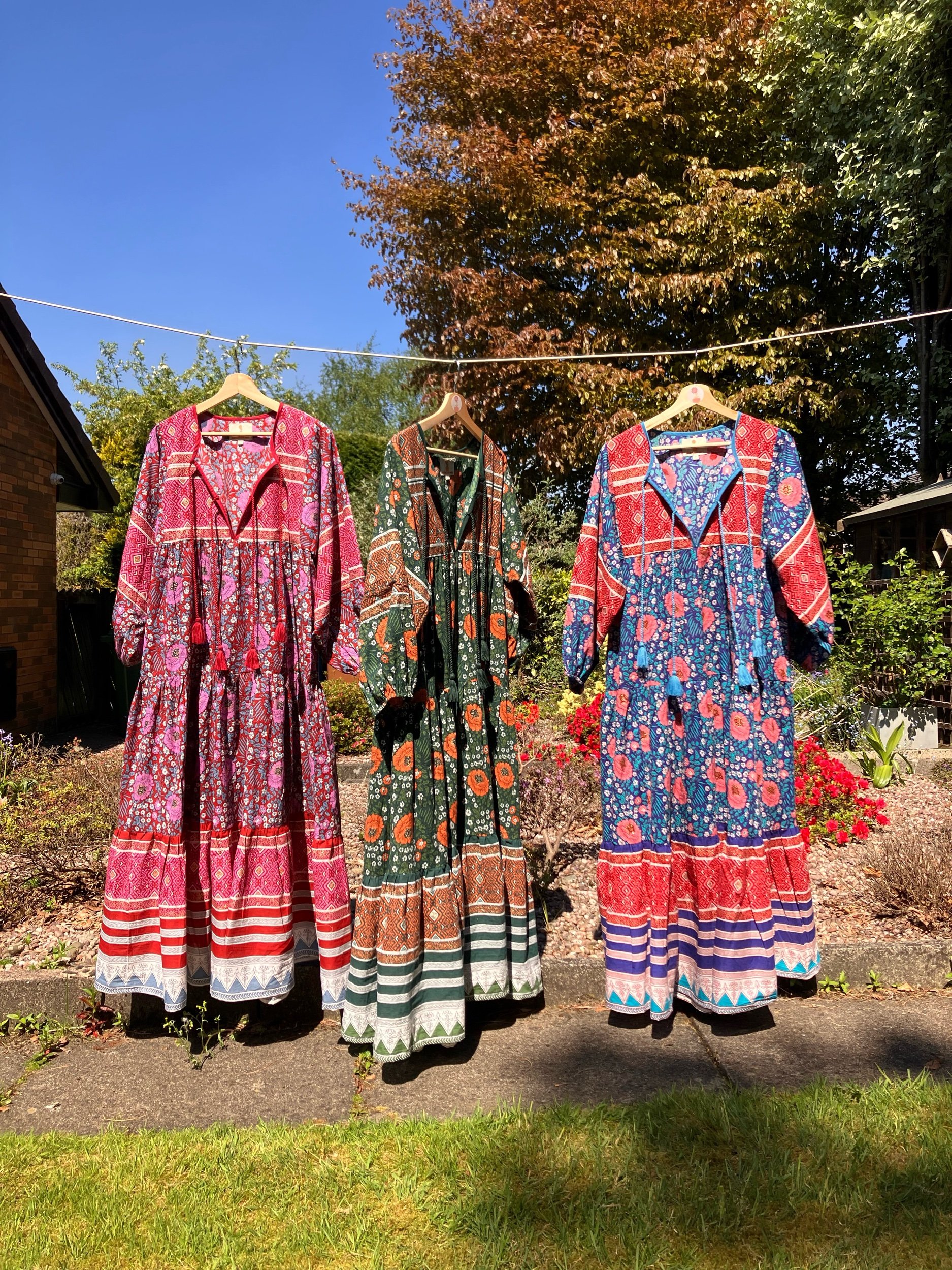 Three colorful bohemian-style dresses hanging on a clothesline in a garden with a blue sky and trees in the background.