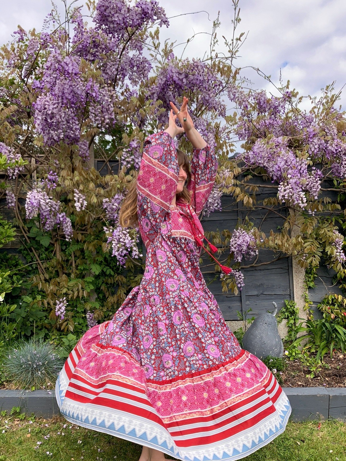 A woman wearing a colorful, floral-patterned dress with red, pink, purple, and white designs, dancing in a garden in front of purple wisteria flowers. She has long brown hair and is smiling.