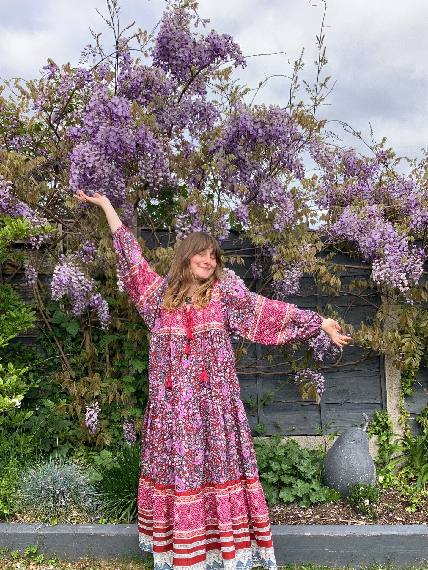Woman in a long, colorful pink dress with floral and geometric patterns standing outdoors with arms raised, smiling, in front of a vine with purple flowers and a black wooden fence, cloudy sky in the background.