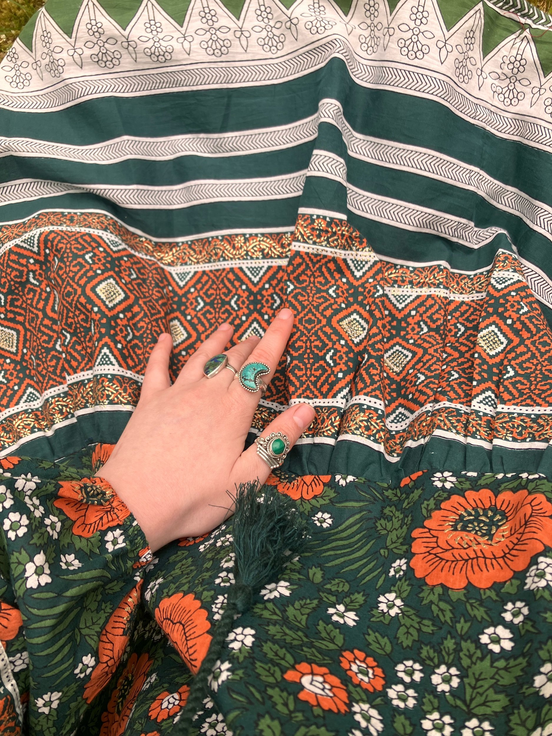 A person's hand with multiple rings resting on a patchwork fabric with floral and geometric patterns in green, orange, white, and black colors.