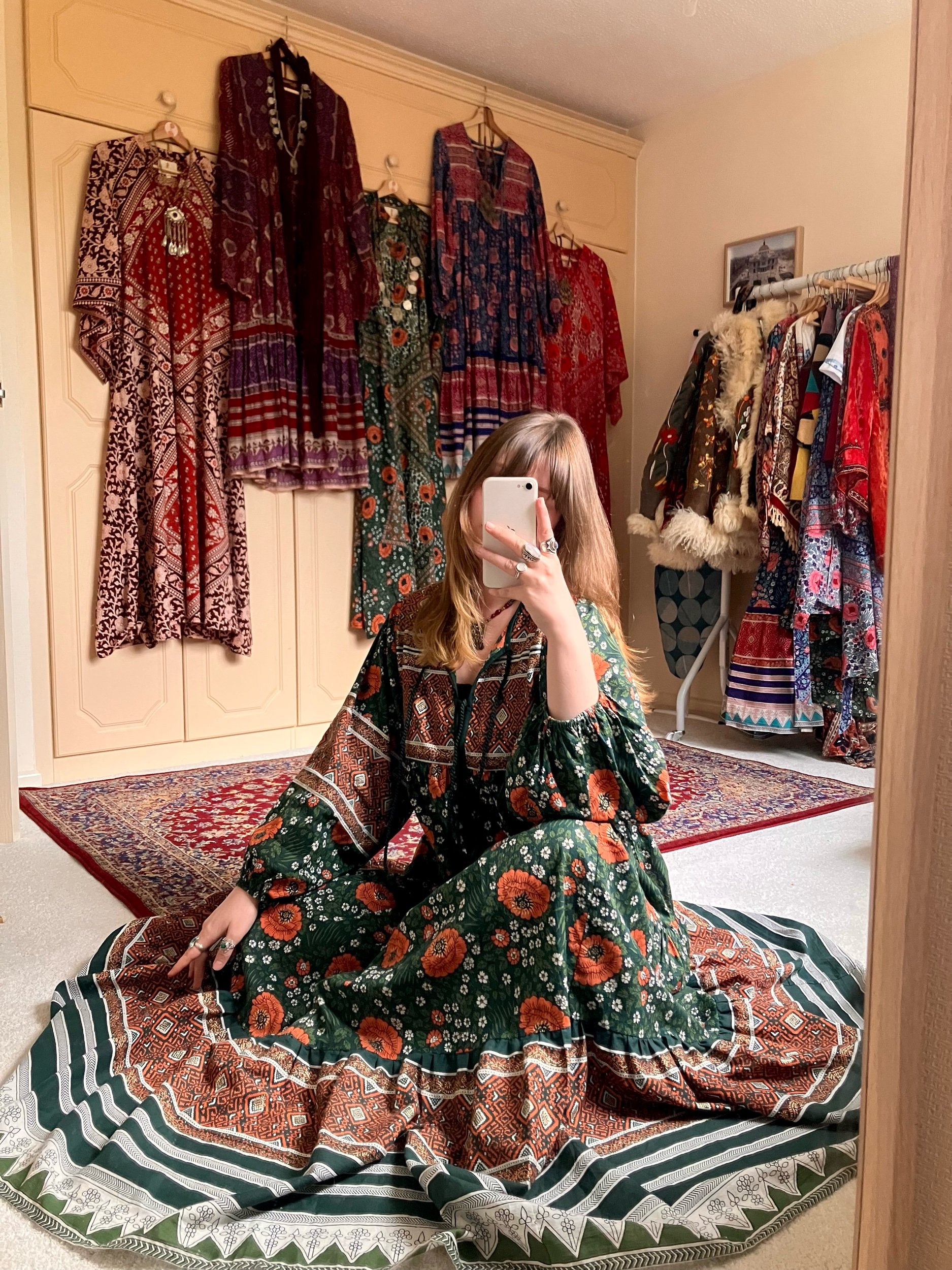 A woman taking a mirror selfie in a room with colorful bohemian-style dresses hanging on the wall and a rack of garments. She is sitting on a patterned blanket on the floor, wearing a long, floral, bohemian dress.