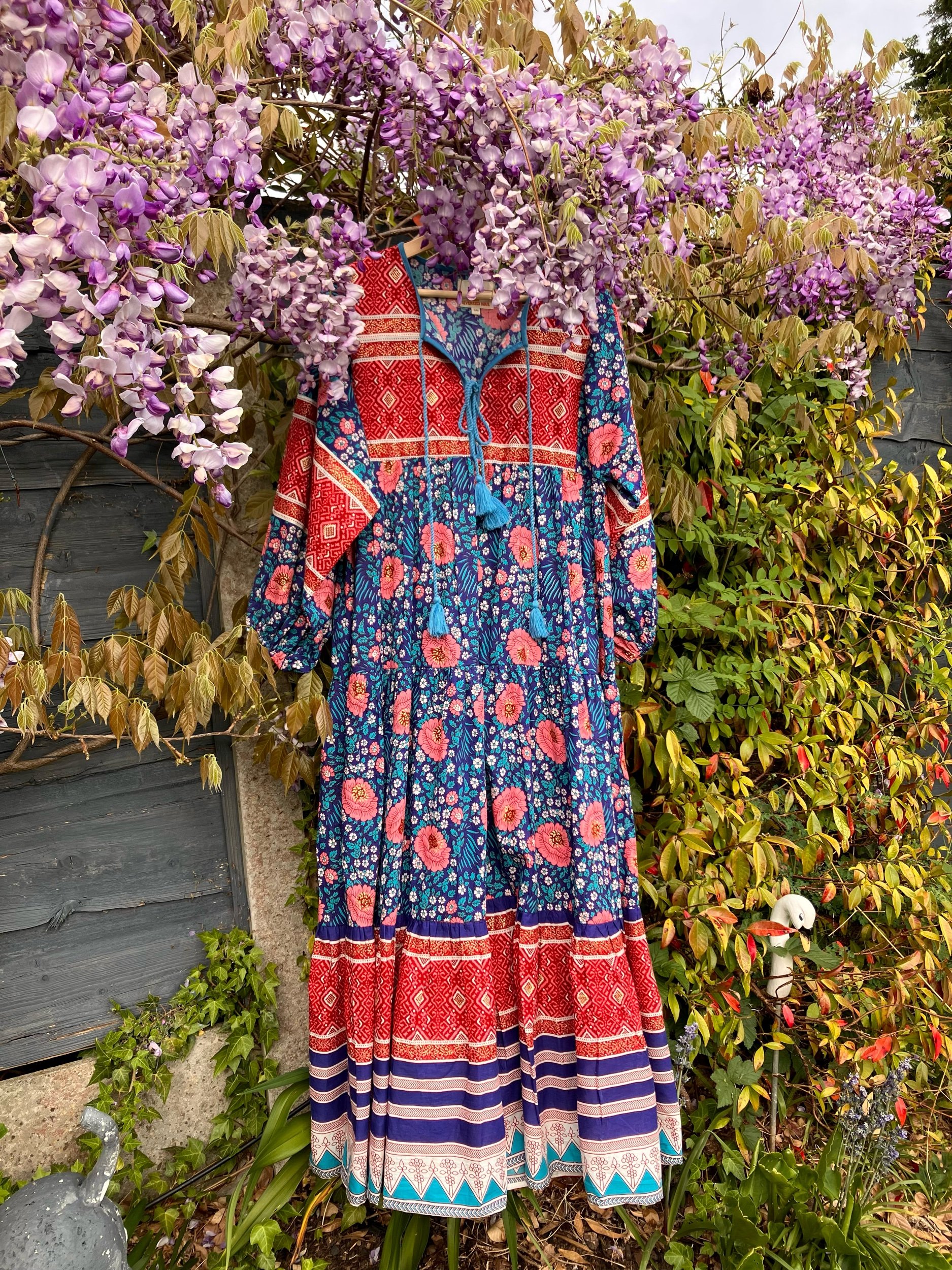 A colorful long dress with floral and geometric patterns hanging on a hanger, surrounded by purple wisteria flowers and green foliage in a garden.