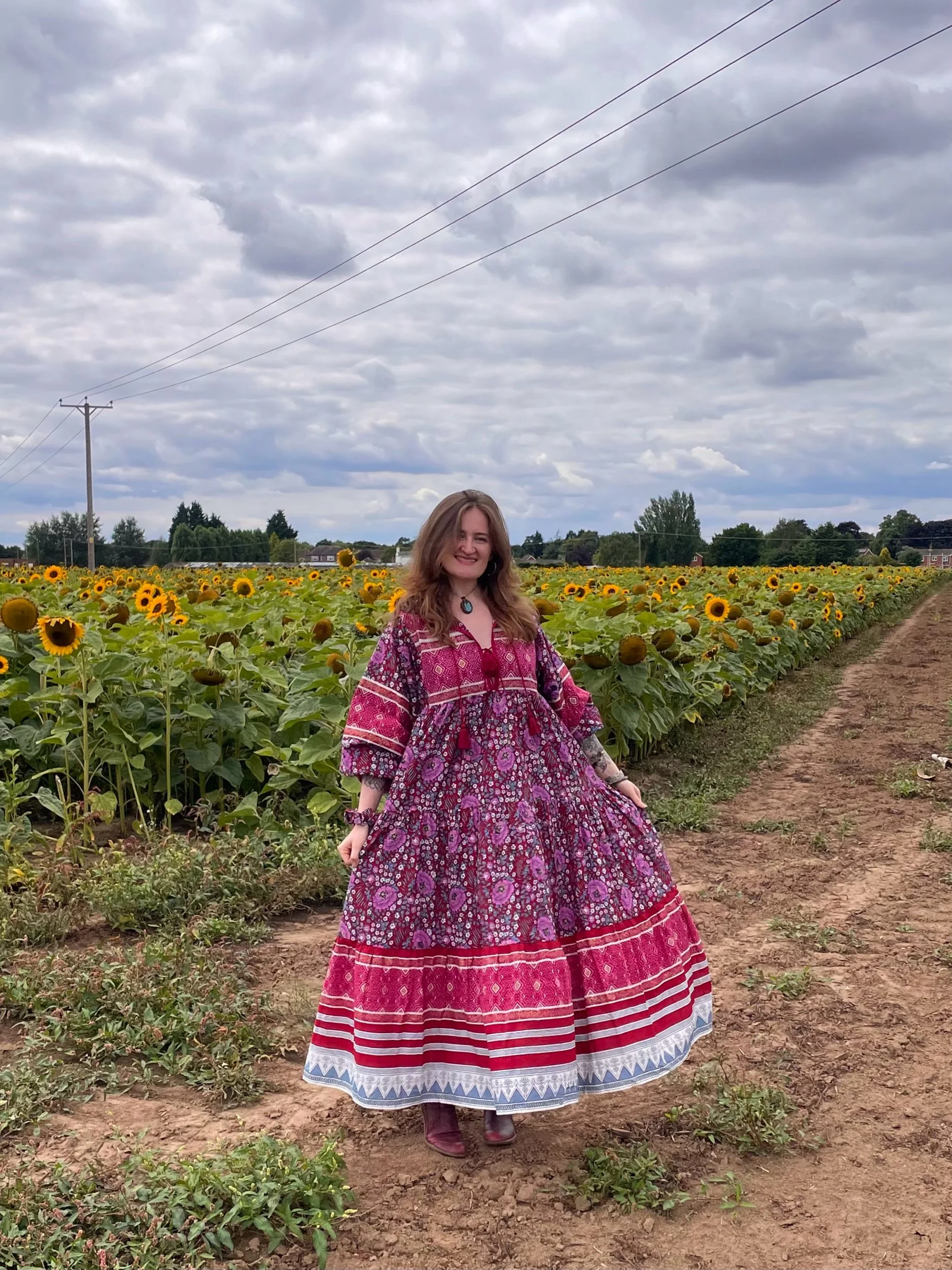 A woman in a colorful, patterned dress standing in a sunflower field under a cloudy sky.
