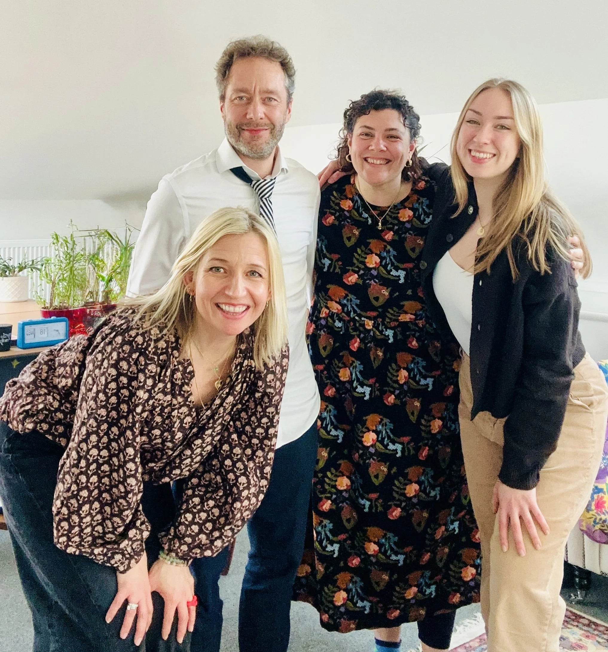 Woman with glasses in a pink sweater smiling and waving at a smartphone on a stand in a bright room.