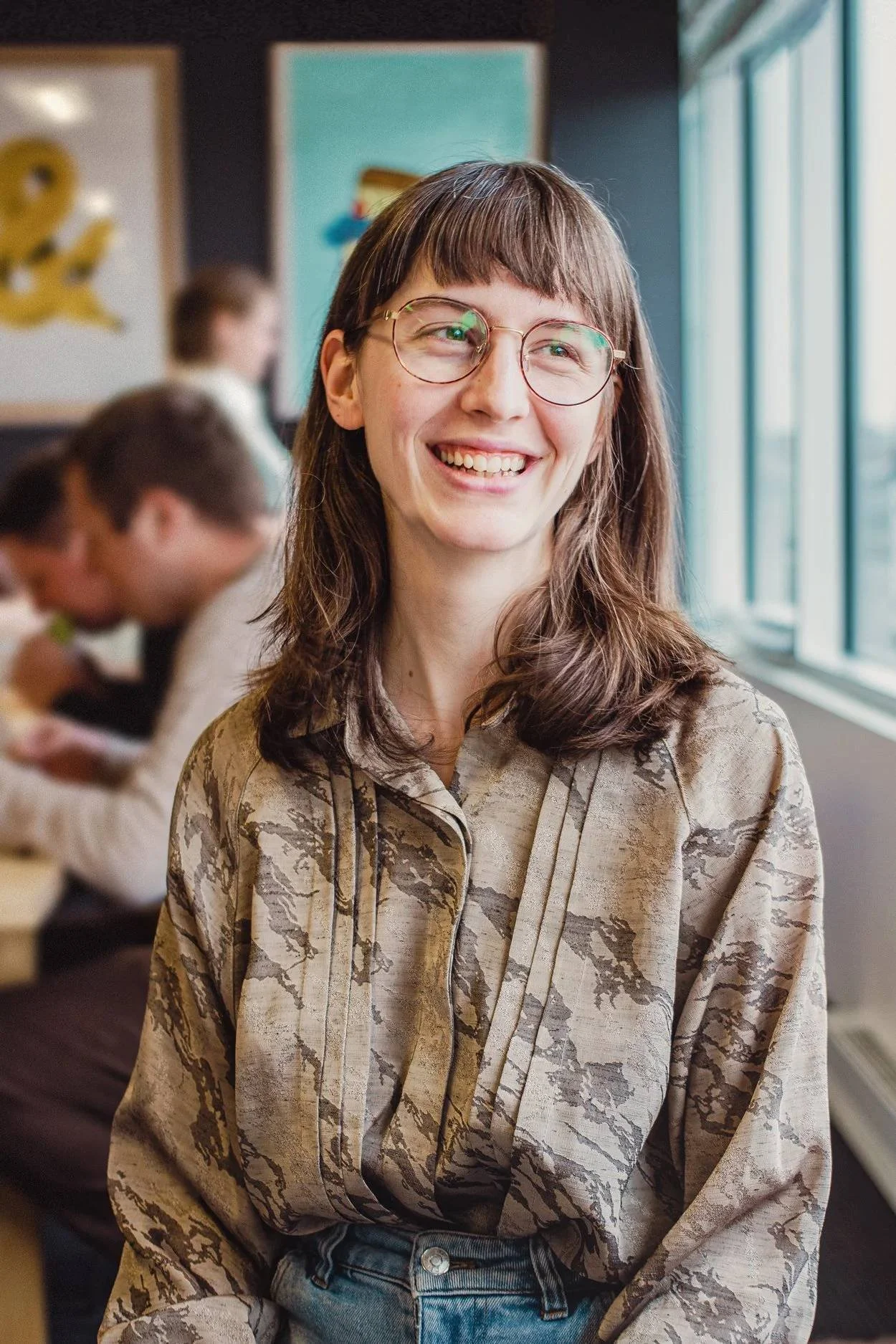 Jeune femme souriante avec des lunettes, portant une chemise à motif beige et des jeans, dans un espace intérieur lumineux avec des œuvres d'art en arrière-plan.