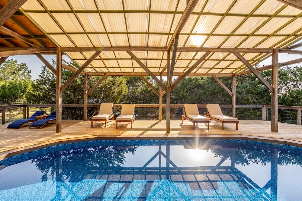 Covered outdoor pool surrounded by large comfortable lounge chairs, creating a relaxing retreat space in a natural Texas setting.
