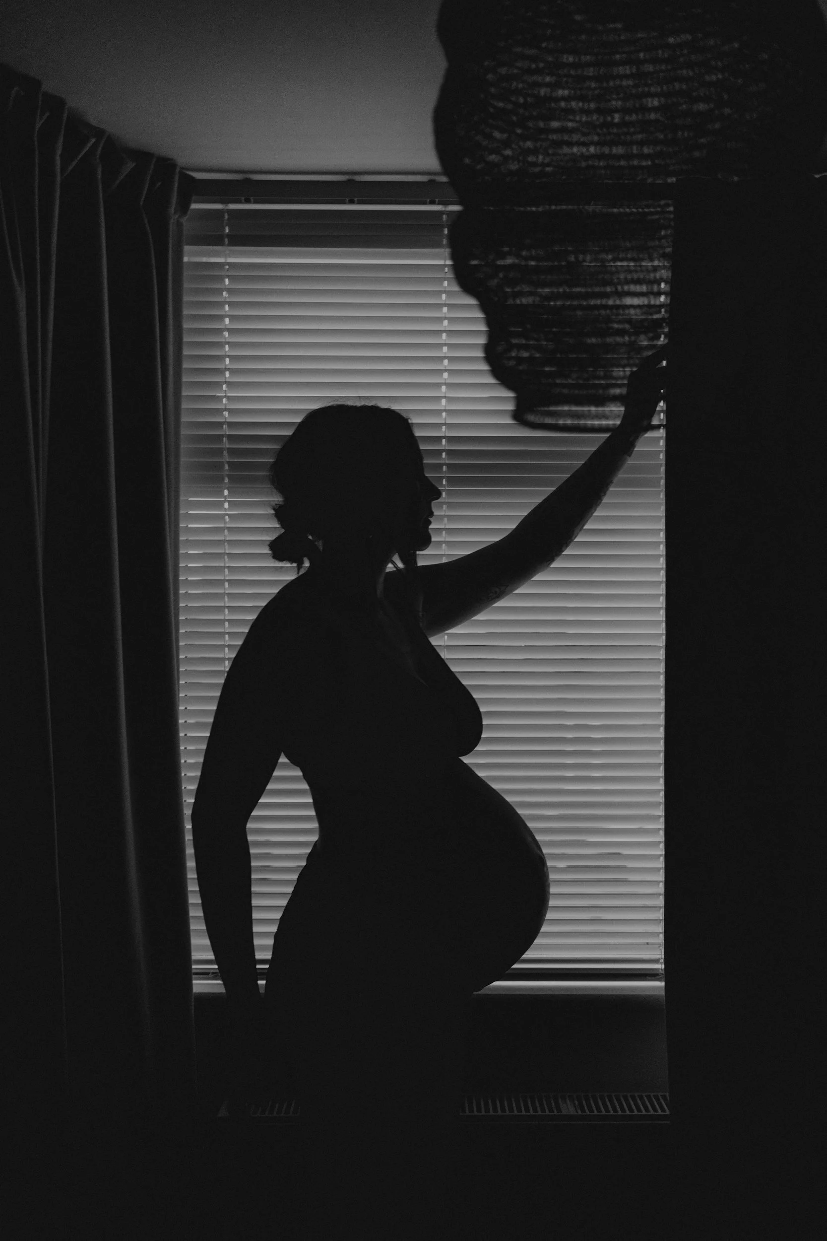Silhouette of a pregnant woman with her hand on her belly, standing against a background of window blinds with curtains, in a dark room.