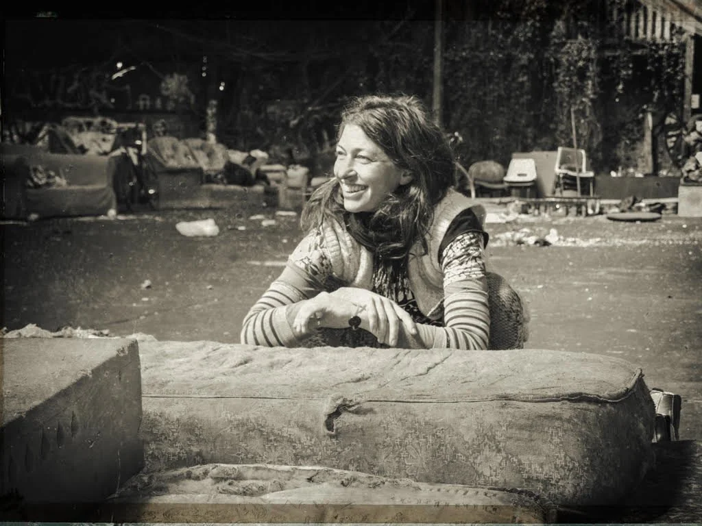 A woman with curly hair and a striped shirt smiling with her arms crossed, sitting behind a large piece of wood, in an outdoor setting, in black and white.