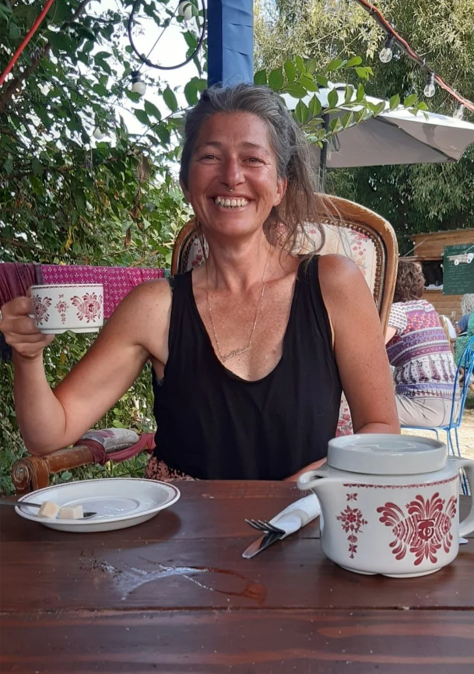 A woman smiling at an outdoor cafe, holding a cup. She is seated at a wooden table with a teapot, a plate with sugar cubes, and utensils. Green foliage and string lights are visible in the background.