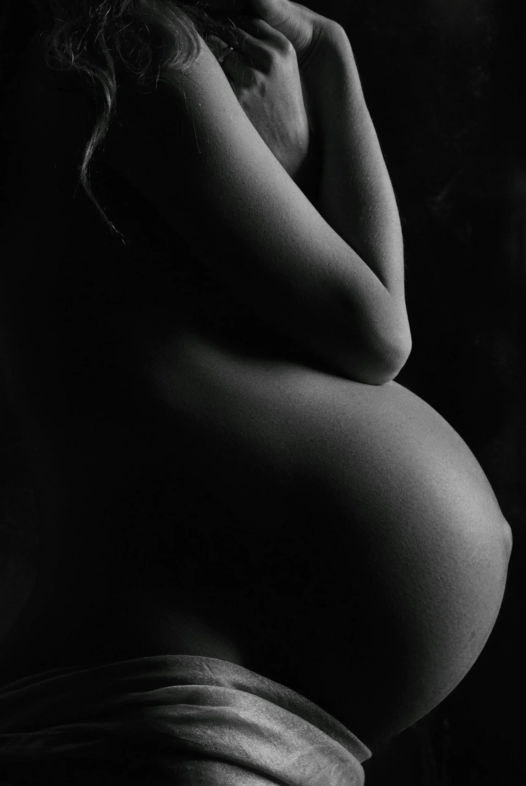 Close-up black-and-white pregnant person with arms crossed over their chest, exposing parts of their face and shoulders, with soft lighting highlighting skin texture and contours.