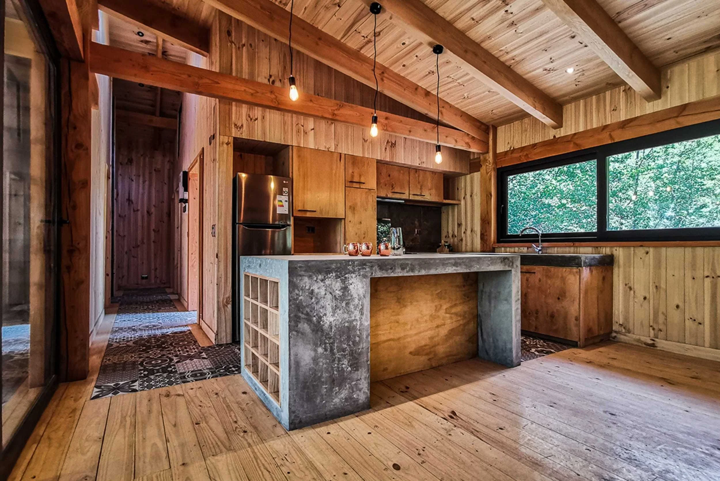 Cocina de madera con techo de vigas expuestas, isla de cemento, lámparas colgantes, refrigerador de acero inoxidable y ventanas grandes con vista al exterior.
