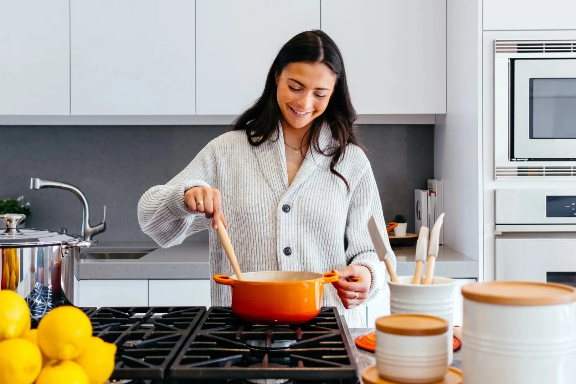 A woman cooking in a modern kitchen, stirring a pot on the stove. The kitchen counter has lemons, containers, and fresh greens, with microwave and kitchen cabinets in the background.