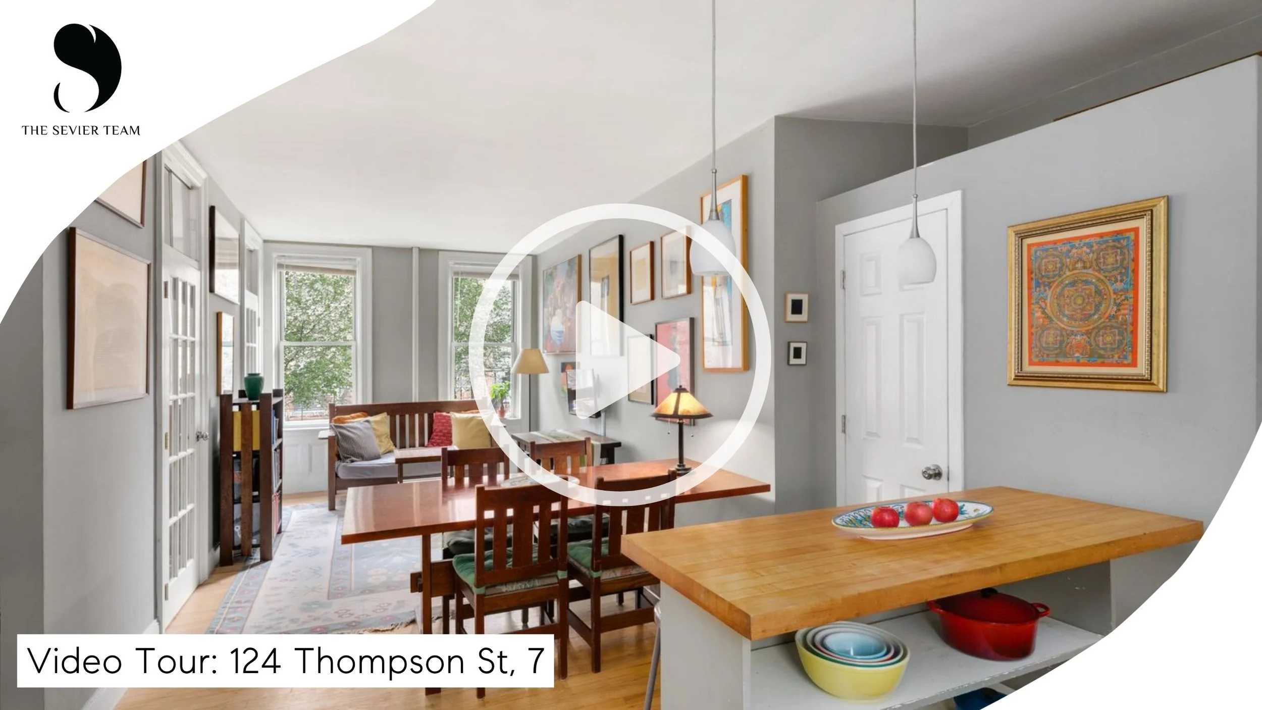 Living room and dining area in a house with multiple windows, framed artwork on the walls, a dining table with chairs, and a kitchen counter with bowls and a red pot.